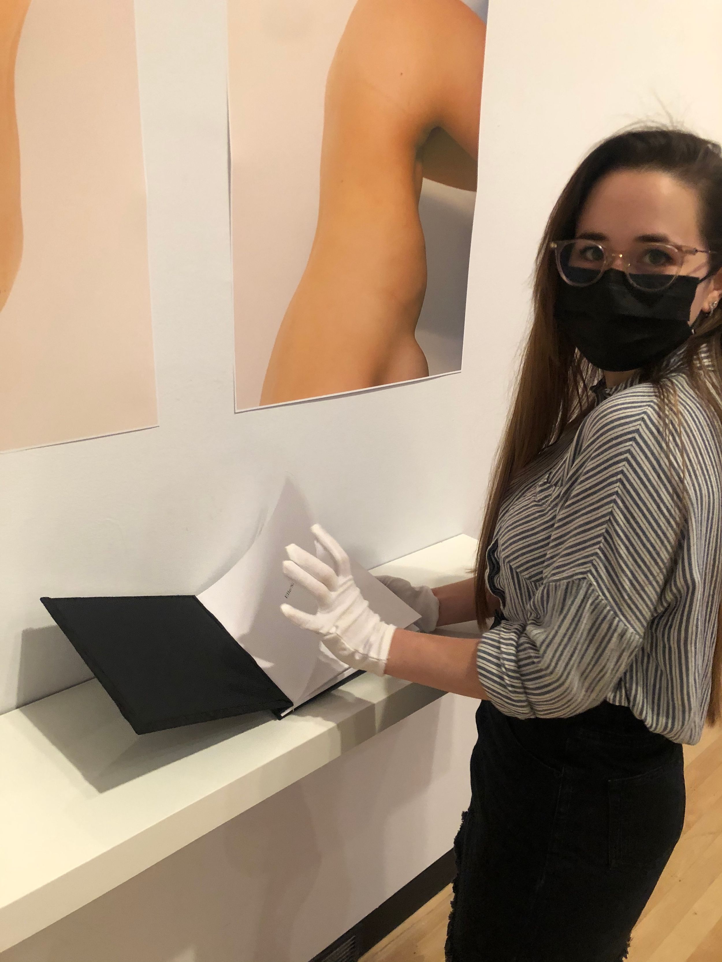 Woman in white gloves and mask opening a black portfolio book at an art gallery exhibition.