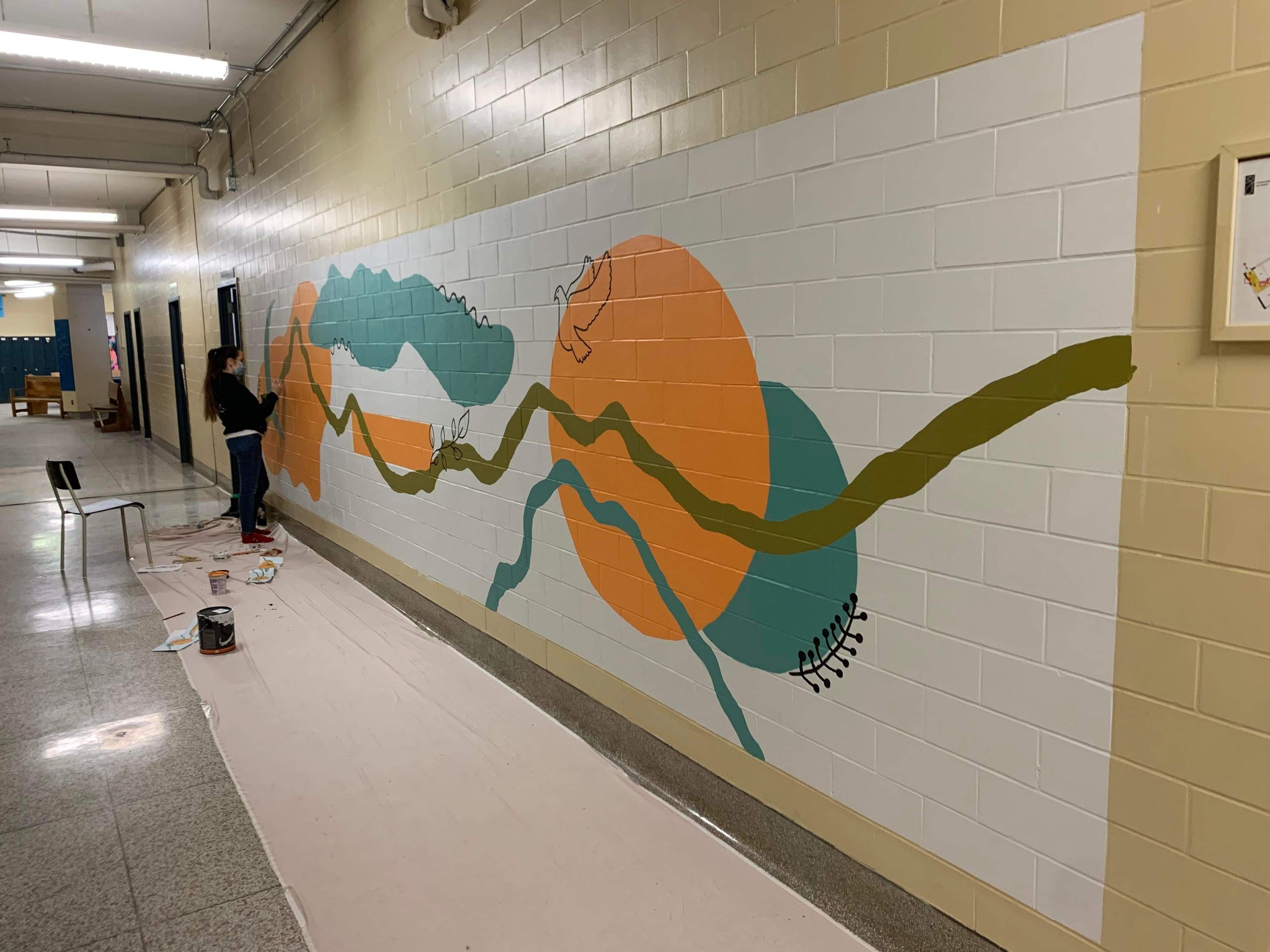 Artist painting a colorful abstract mural with teal, orange, and olive tones on a school hallway wall.