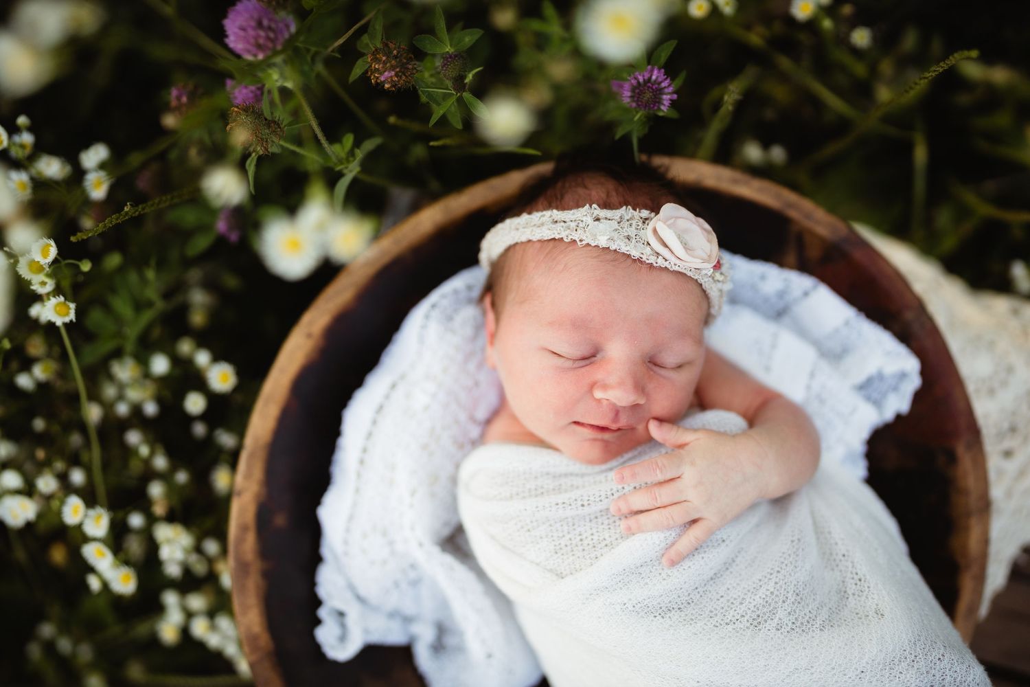 Sleeping newborn wrapped in white lies in wooden bowl surrounded by delicate wildflowers.