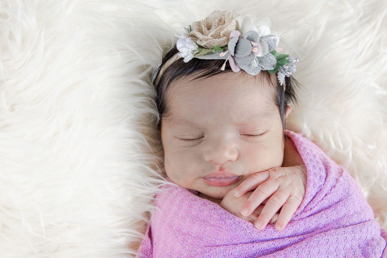 Sleeping newborn wrapped in pink blanket wearing delicate floral headband on white fur backdrop.