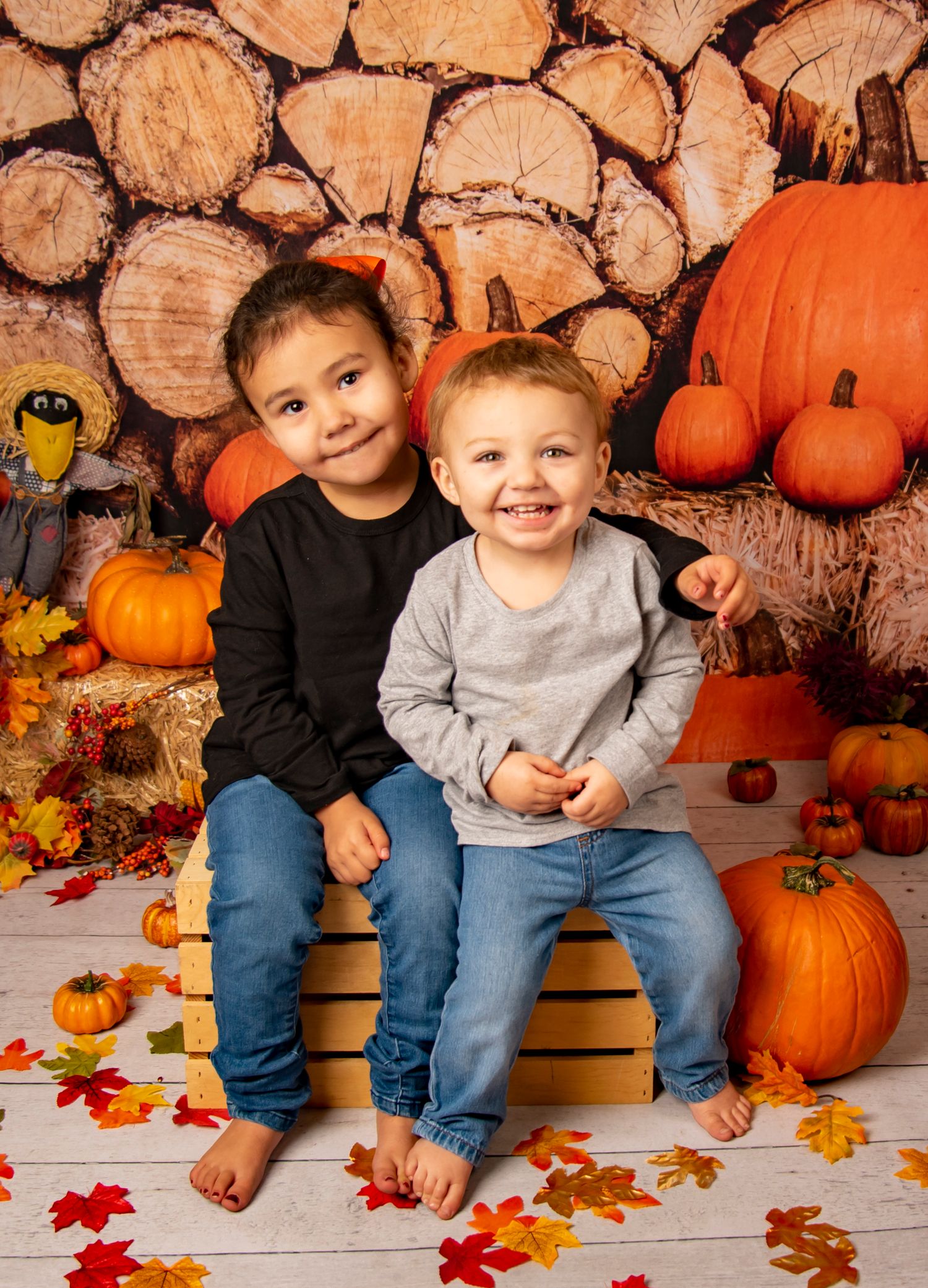 Two children sit on wooden crates surrounded by autumn pumpkins and fall leaves.