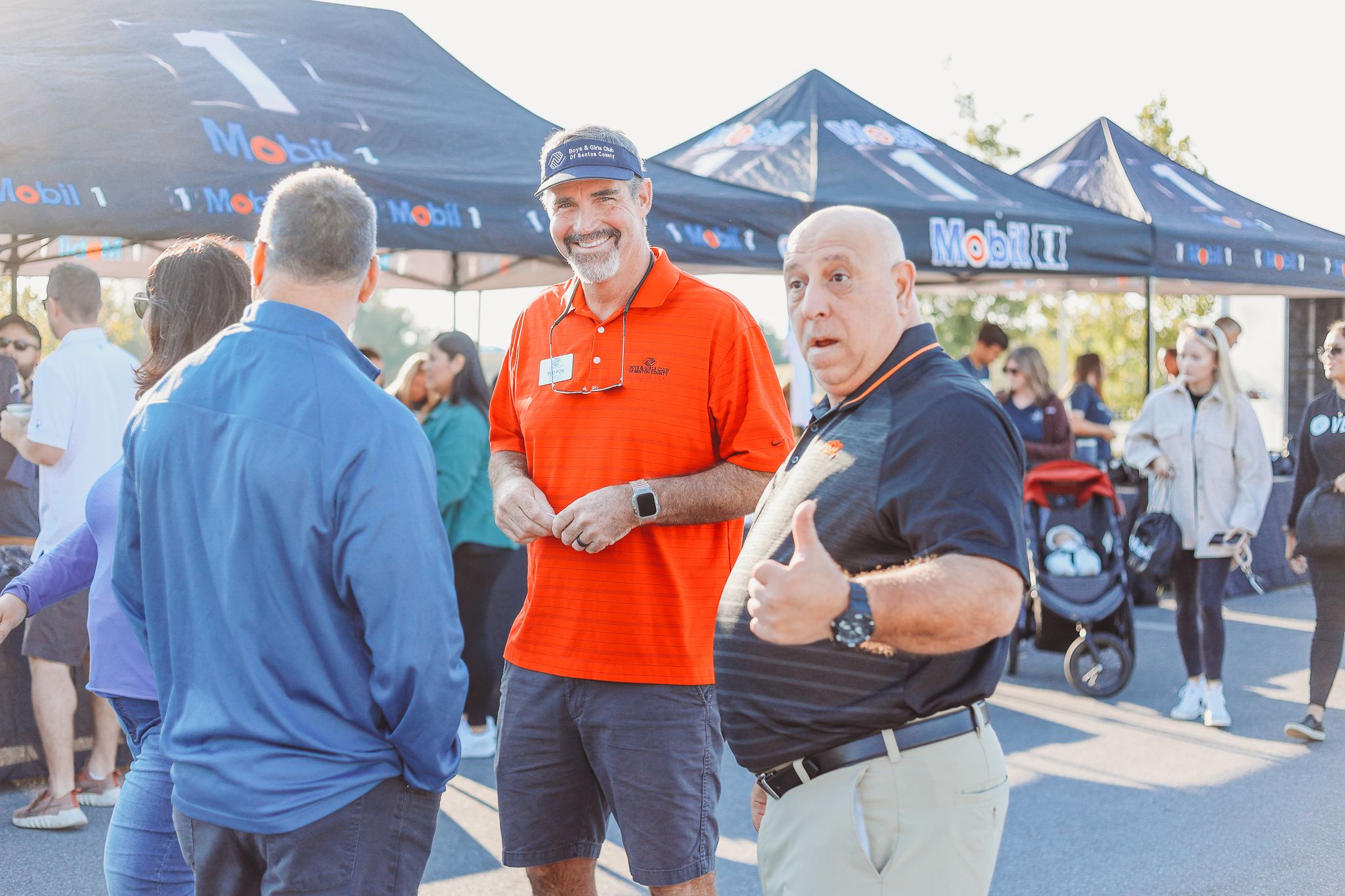 A group of people chatting at an outdoor event with blue pop-up tents visible in the background.