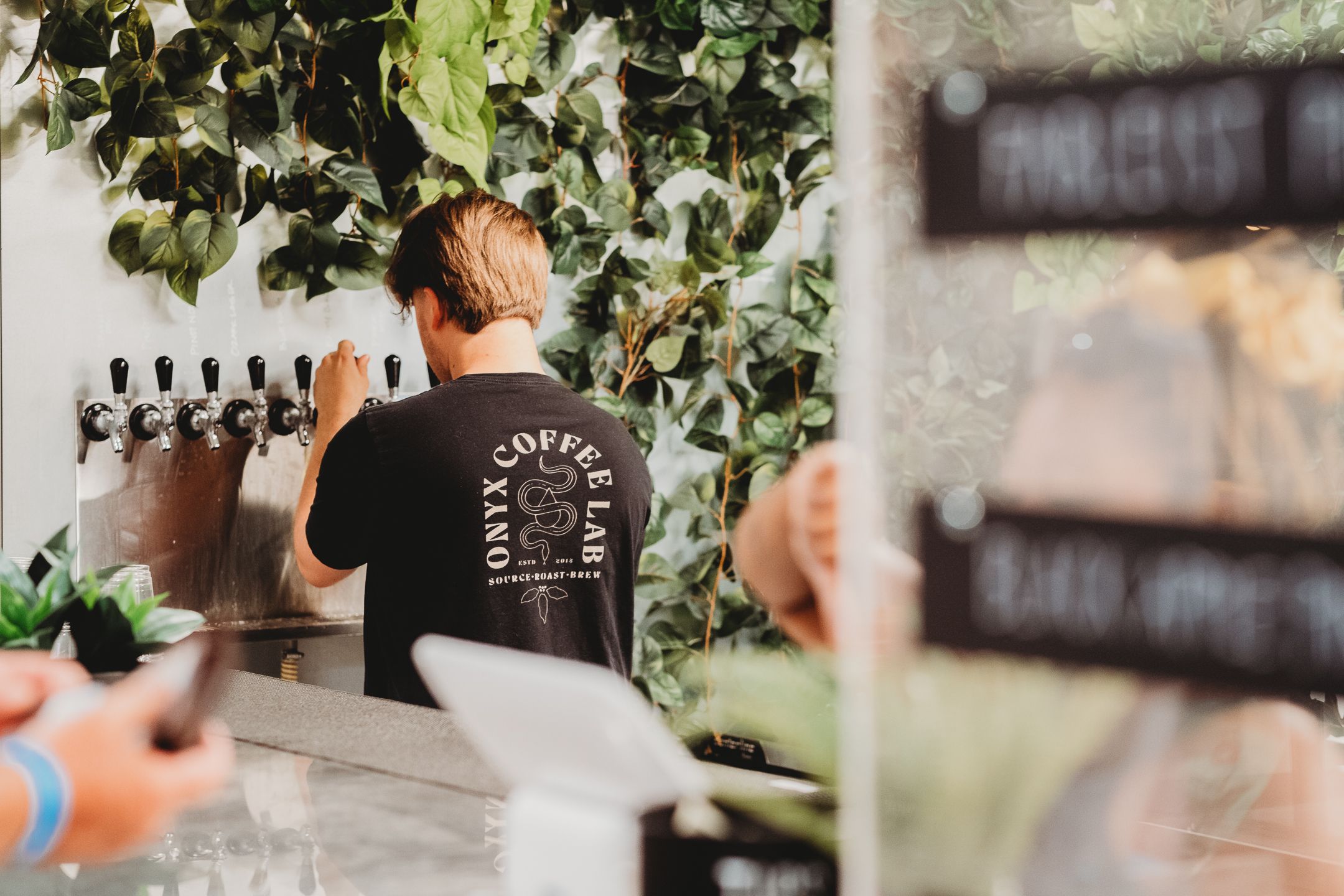 Staff member working behind bar counter with greenery and taps visible in background.