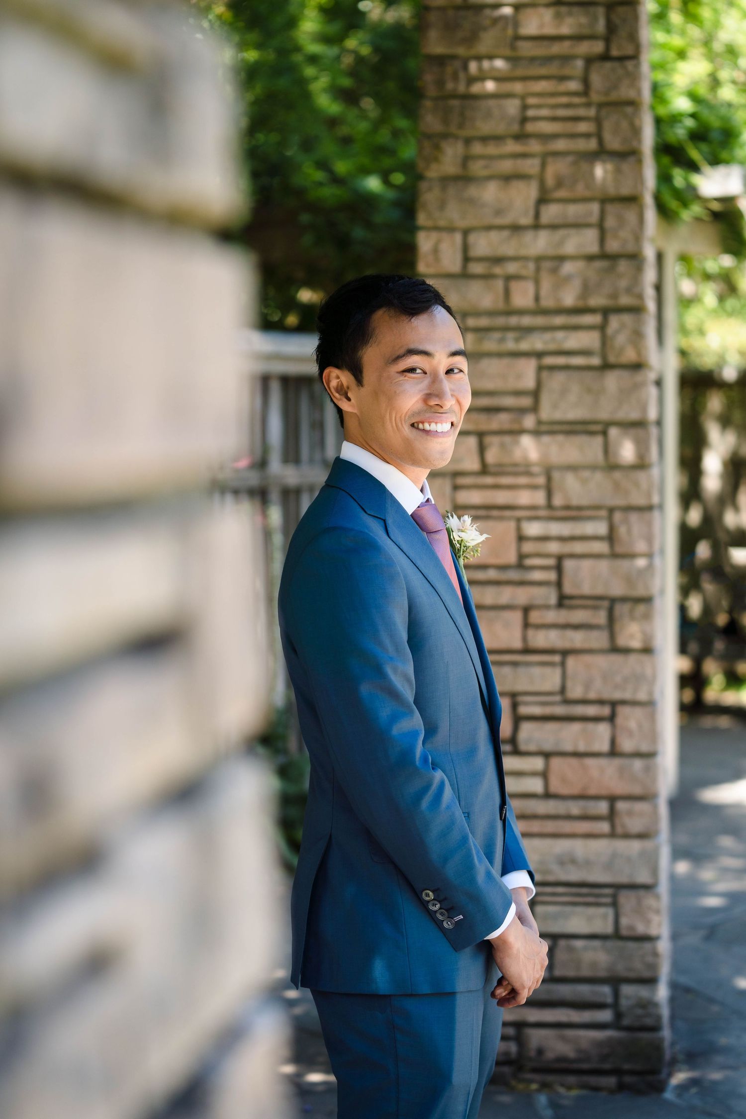 Wedding portrait against stone column with natural summer garden backdrop.