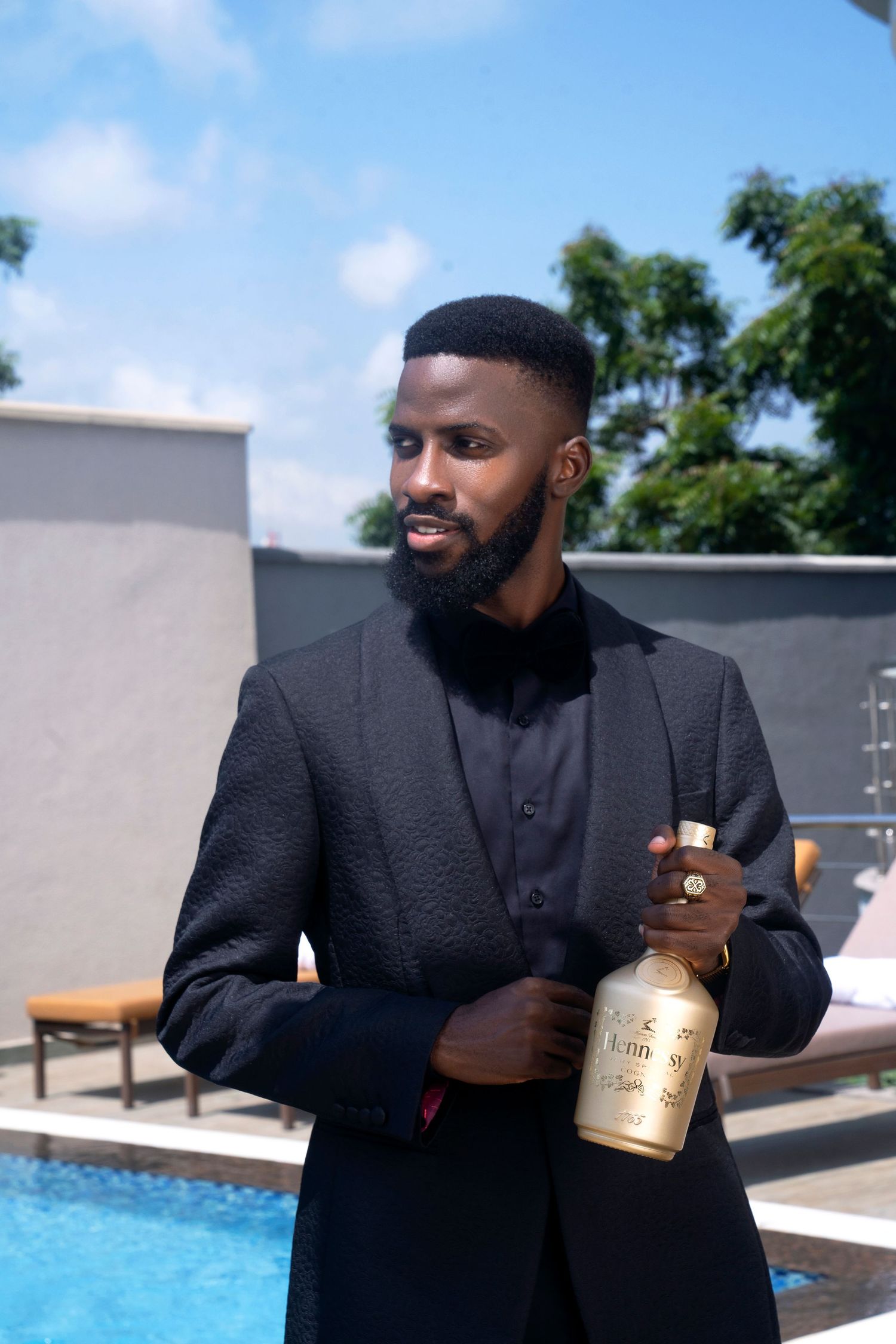 Professional in black suit holds gold bottle by poolside on sunny day.