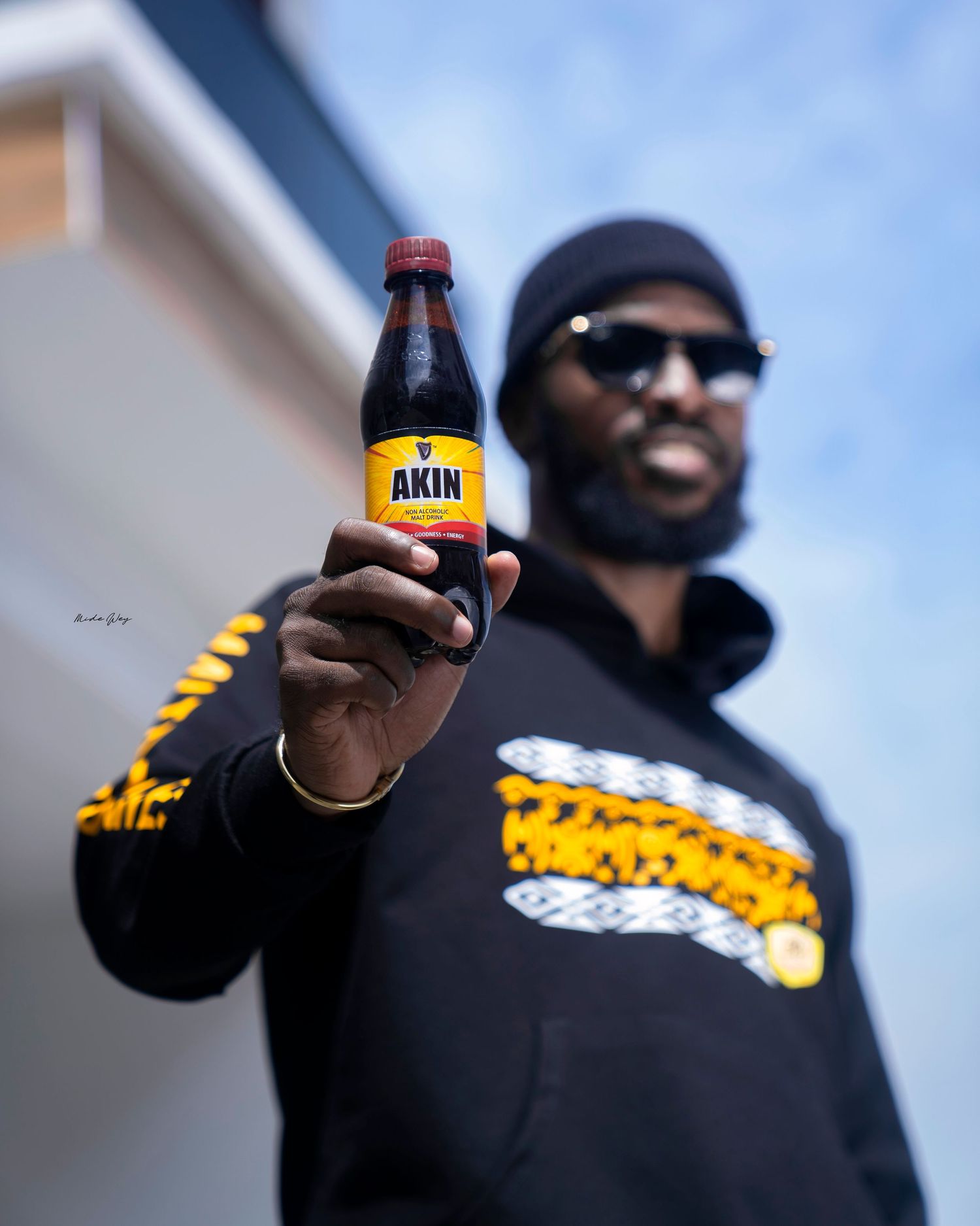 A person in a dark hoodie holds up a bottle of Akin drink against a blue sky.