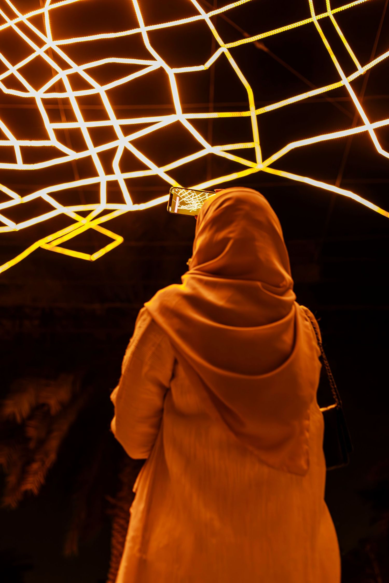 Figure in an orange hijab stands beneath glowing geometric light installations at night.