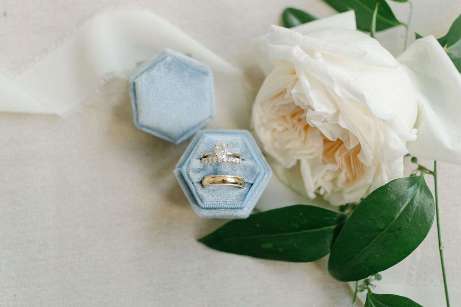 Gold diamond engagement ring and wedding band displayed in blue velvet hexagon ring box with white rose.