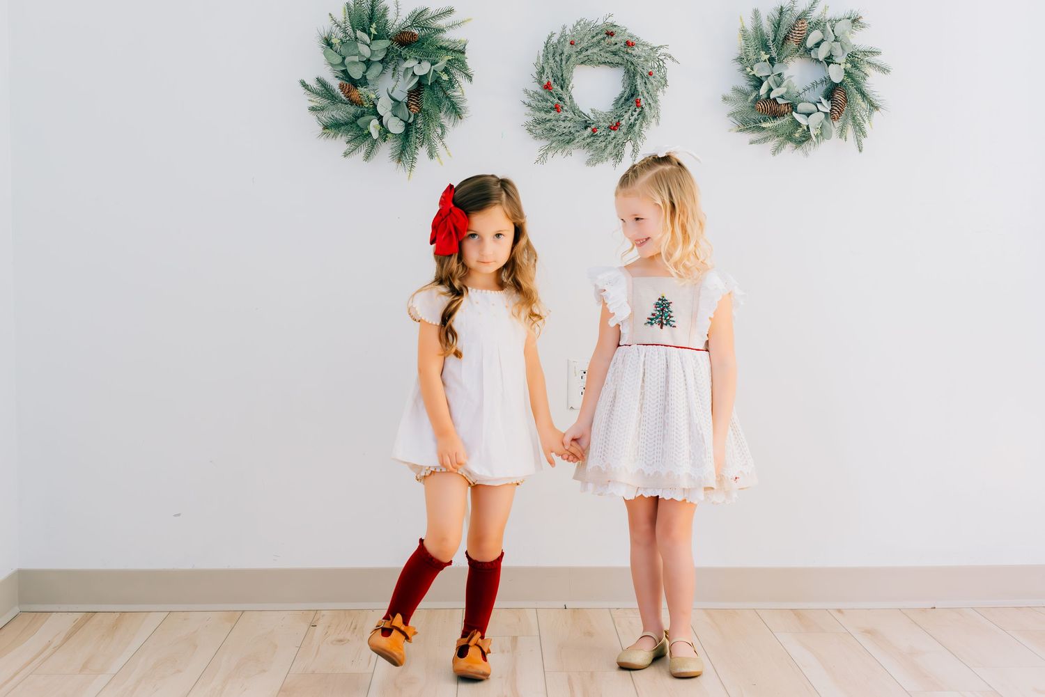 Two young children in light dresses hold hands against a wall decorated with green holiday wreaths.