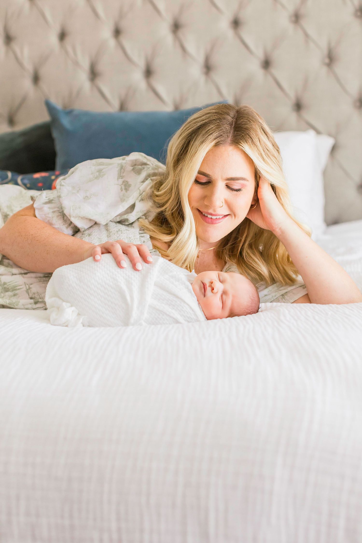 A tender moment between mother and newborn baby lying together on a tufted headboard bed.
