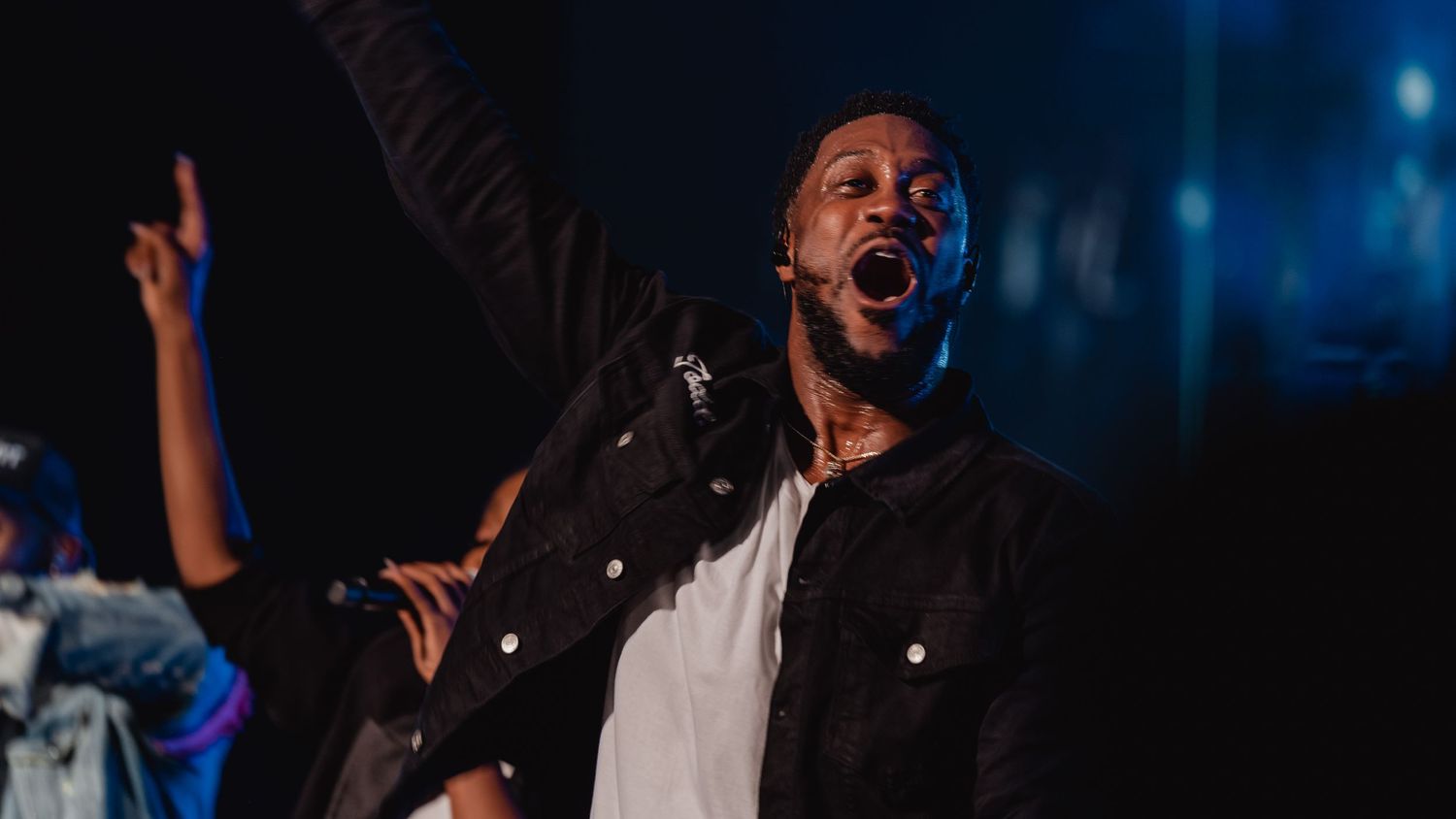 A passionate performer sings on stage with one hand raised, wearing a black jacket under dramatic blue stage lighting.
