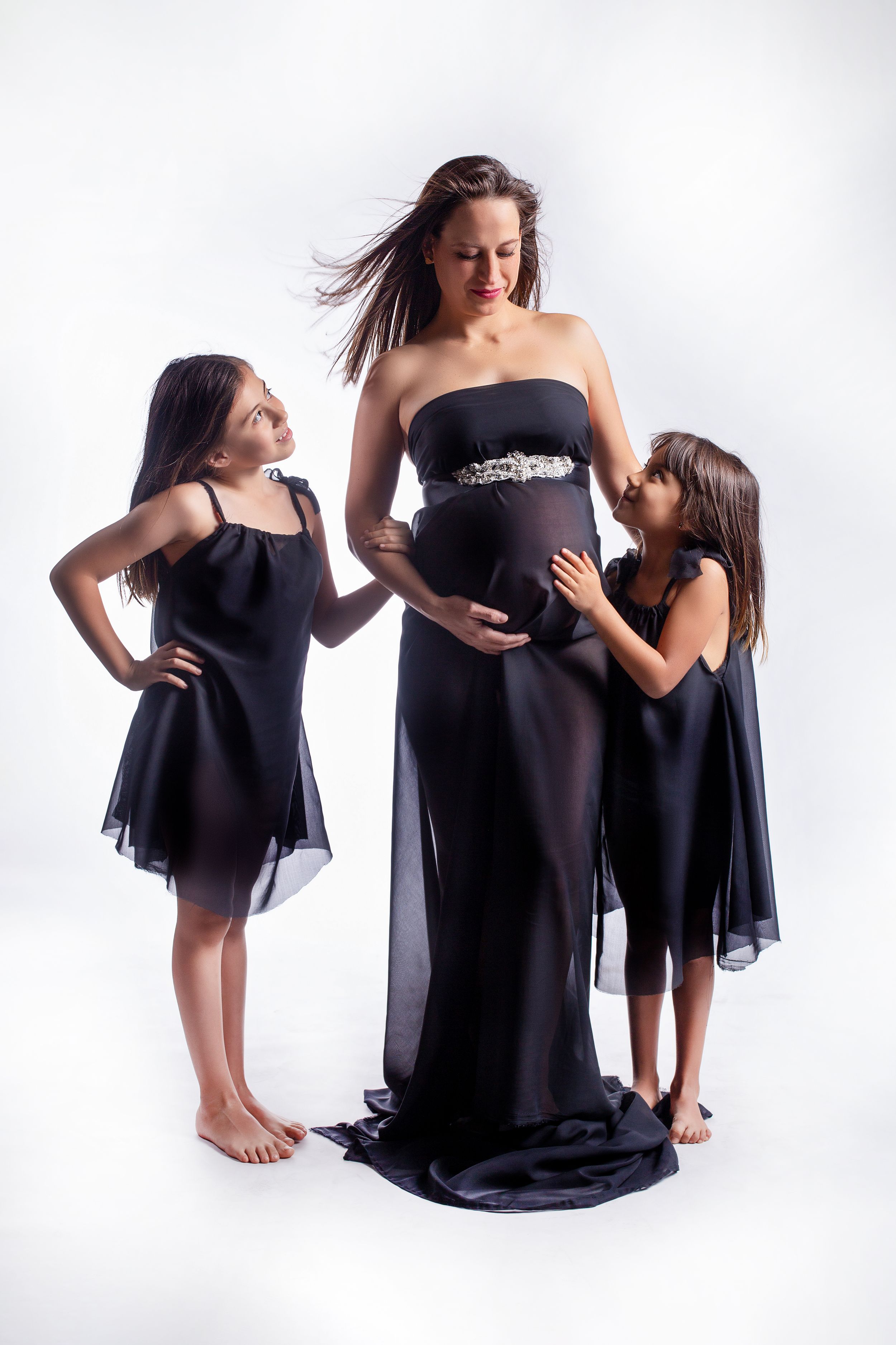 Pregnant woman in black gown with two young girls in black dresses touching her baby bump.