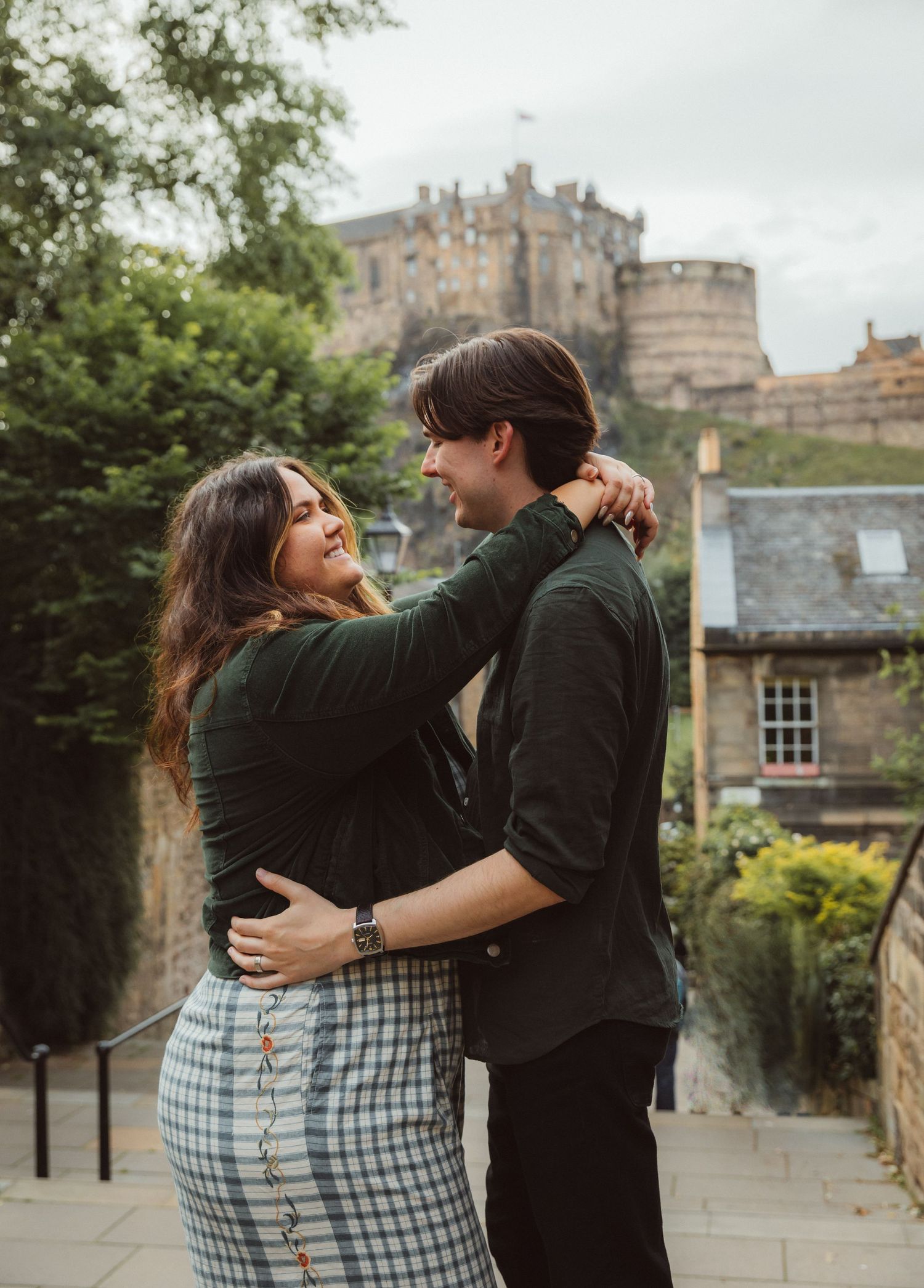 A couple shares an intimate moment in front of Edinburgh Castle on a cloudy day.
