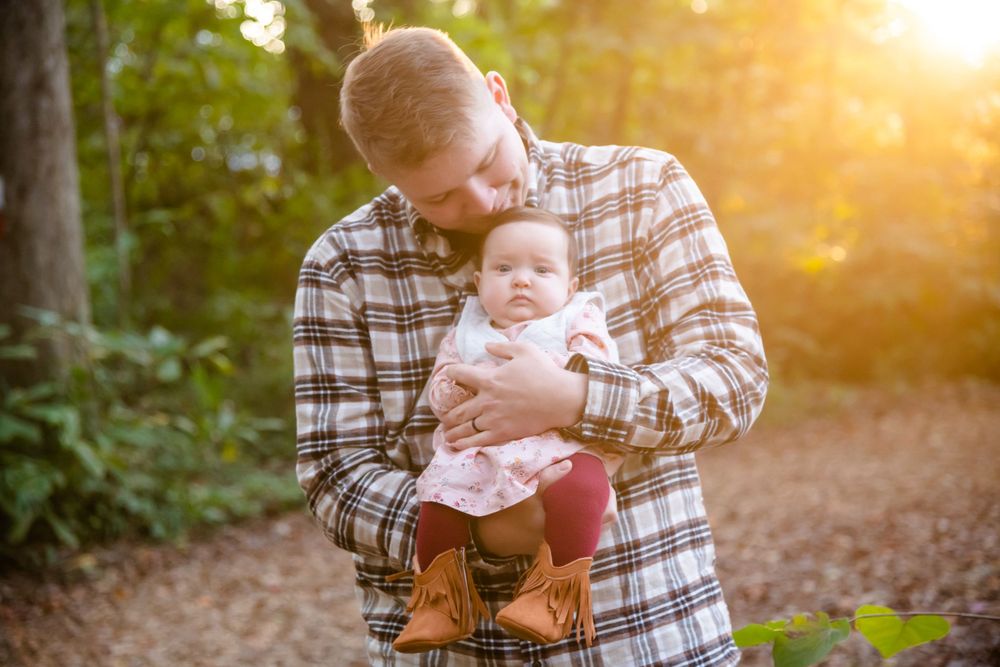 Father in plaid shirt holding baby during golden sunset in wooded area.