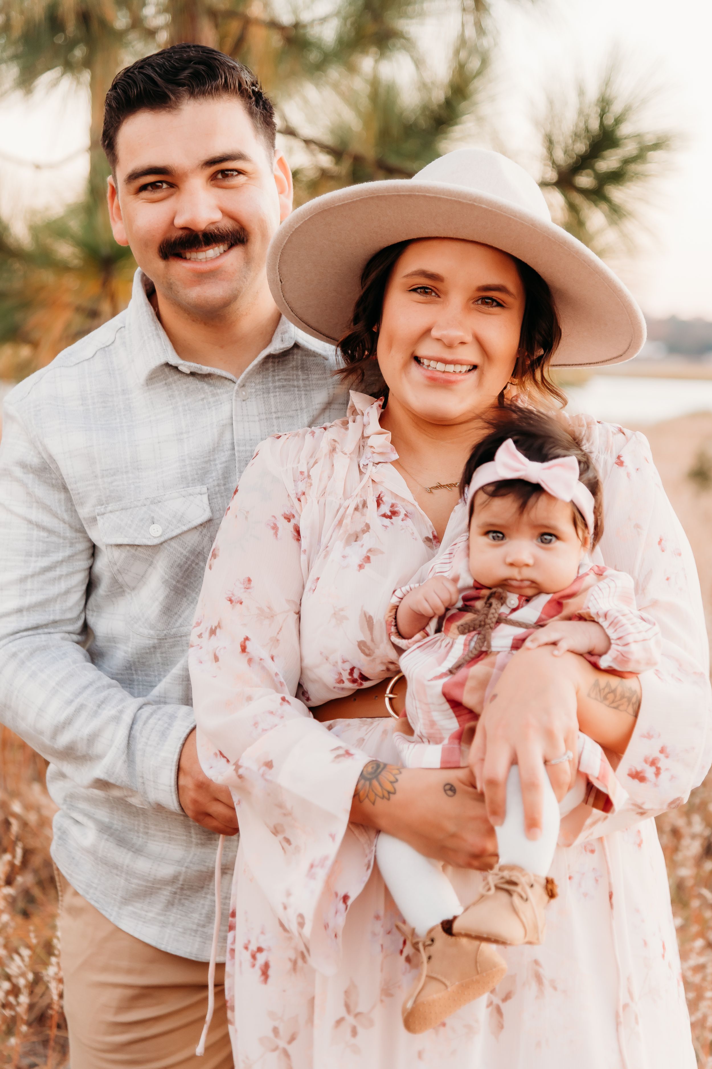 Family standing together outdoors with parent wearing wide brim hat and floral dress holding baby.