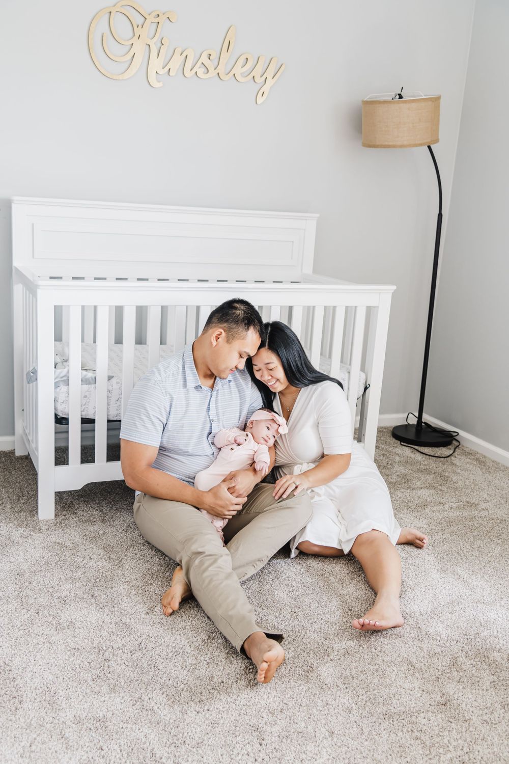 A couple sits on carpet with their newborn baby in front of a white crib with 'Paisley' written on the wall.