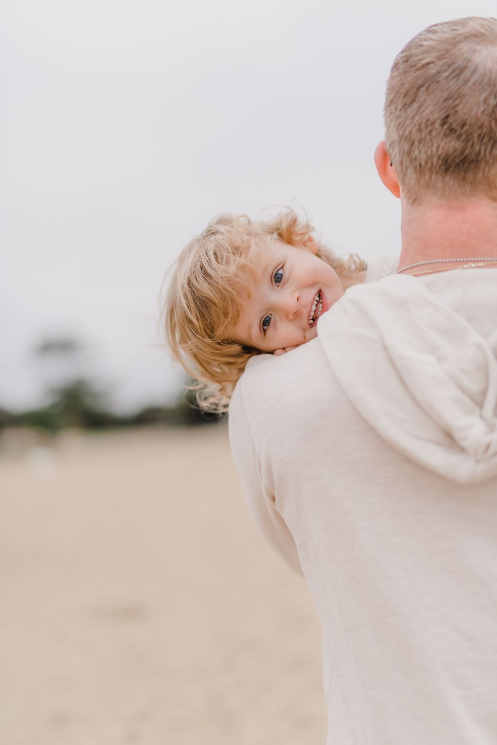 Small child with curly blonde hair peeking over shoulder with playful expression on beach.