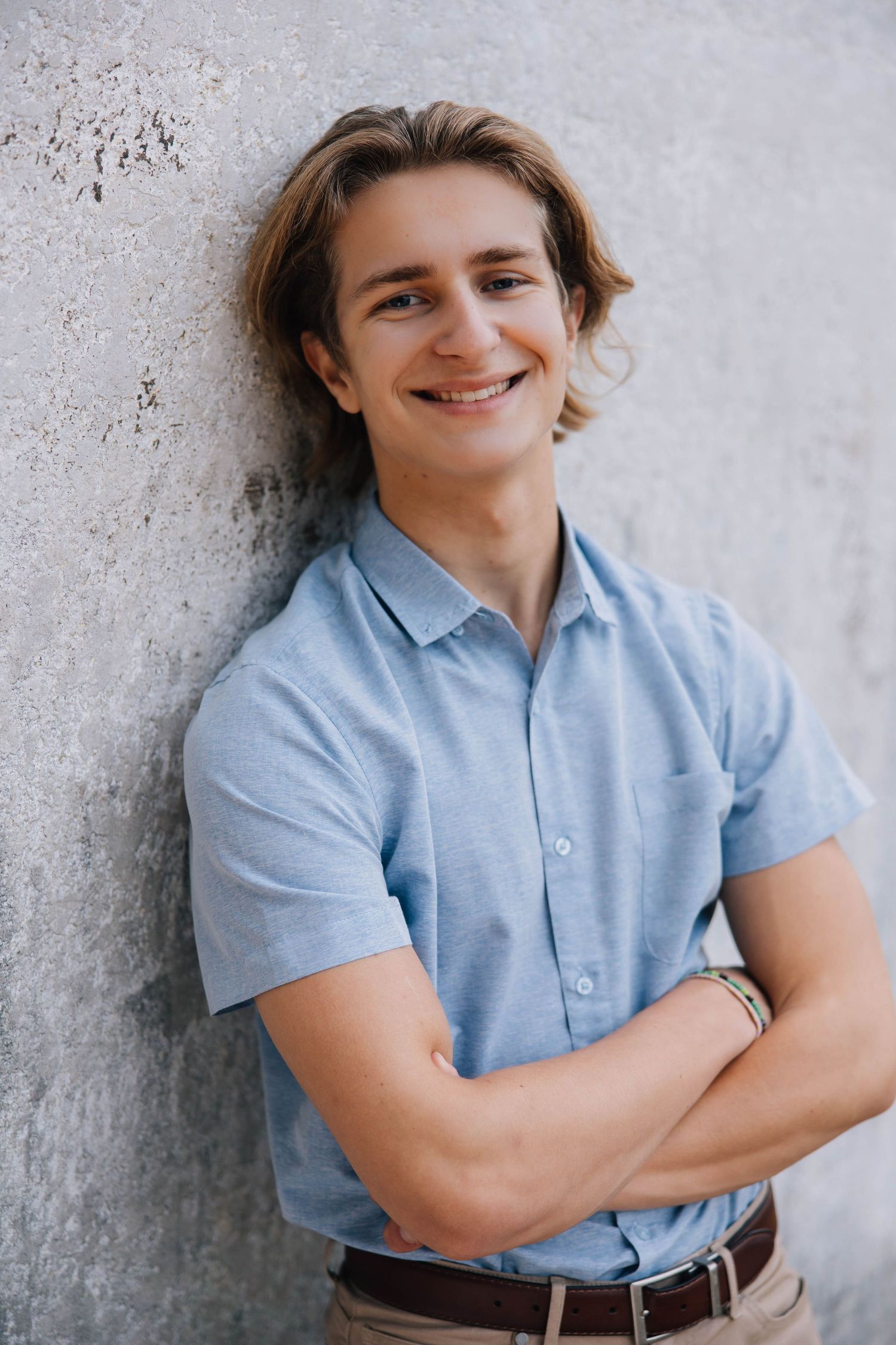 Smiling young man in blue shirt leans against concrete wall with arms crossed.