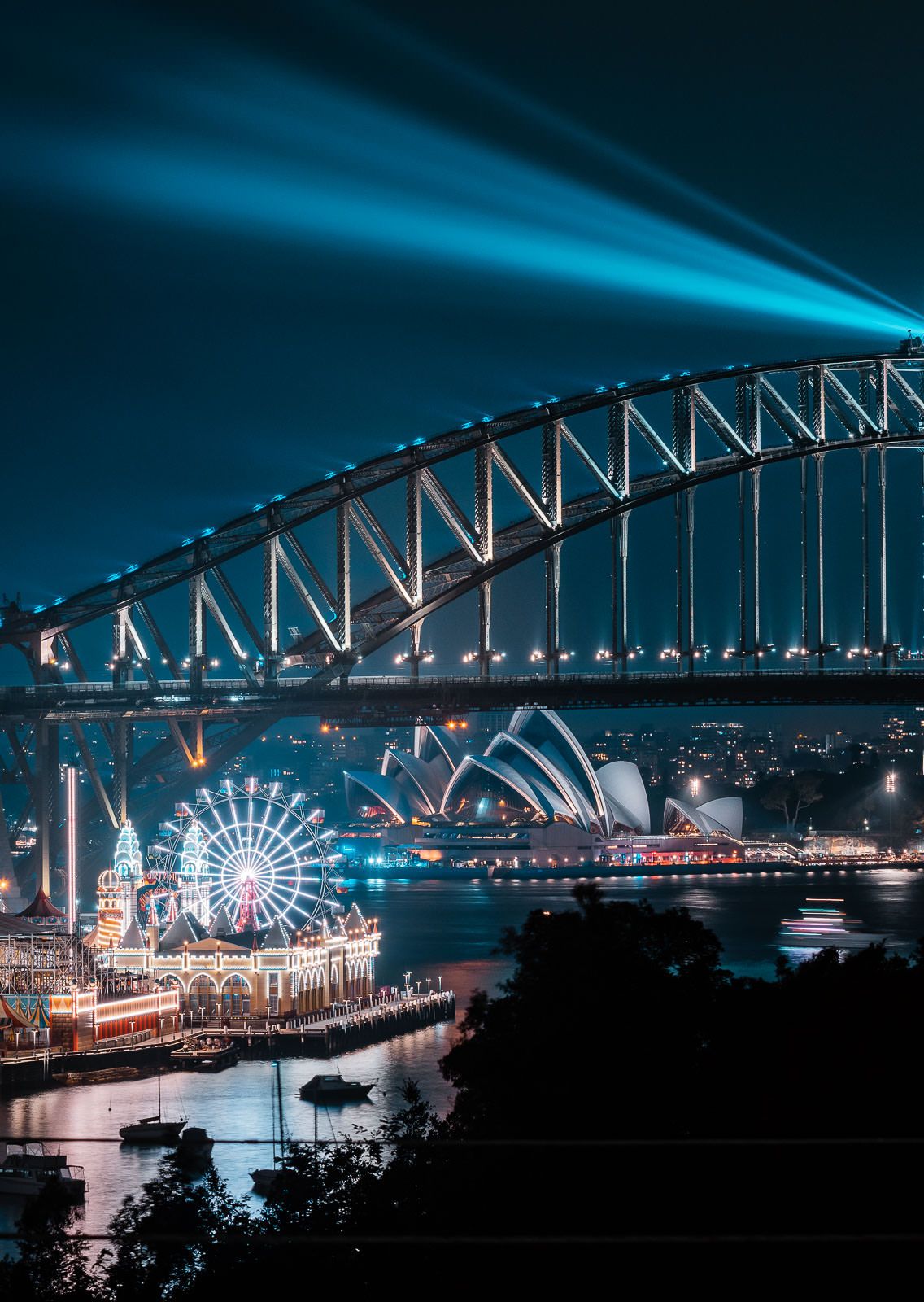 Sydney Harbour Bridge and Opera House illuminated at night with Luna Park ferris wheel visible.