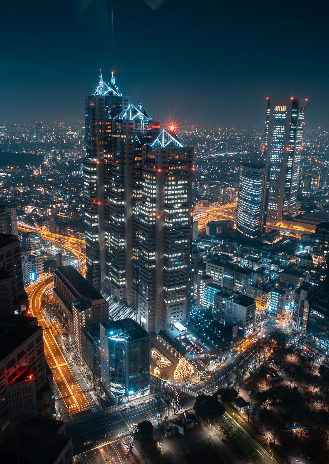 Modern skyscrapers lit up at night in a cityscape with glowing orange highways below.