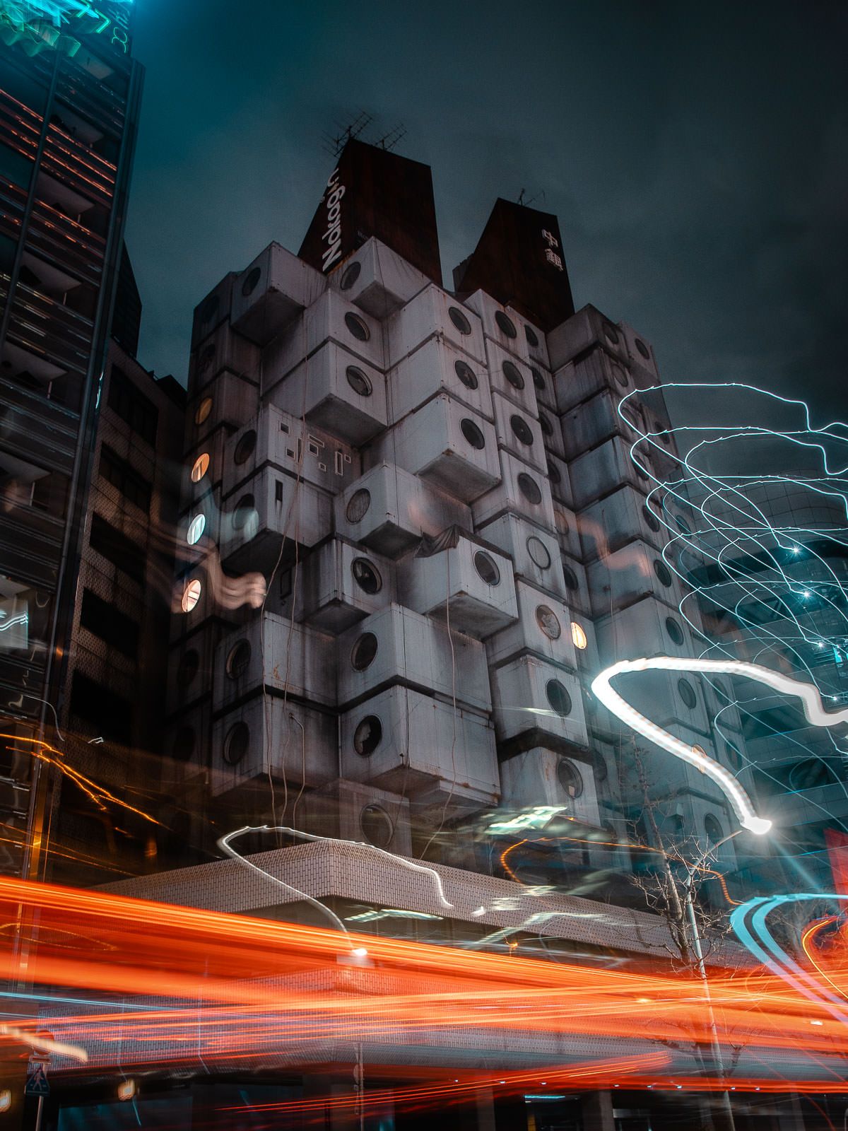Nakagin Capsule Tower in Tokyo with light trails from traffic below against a moody blue sky.