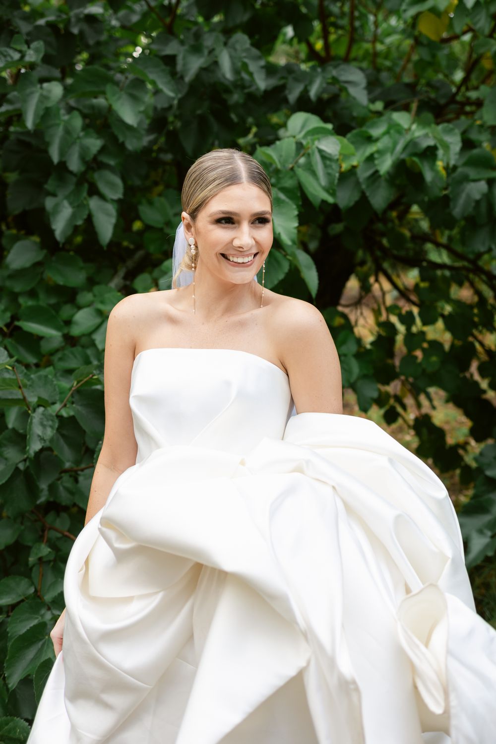 Beautiful bride in strapless white ball gown poses outdoors against lush green foliage background.