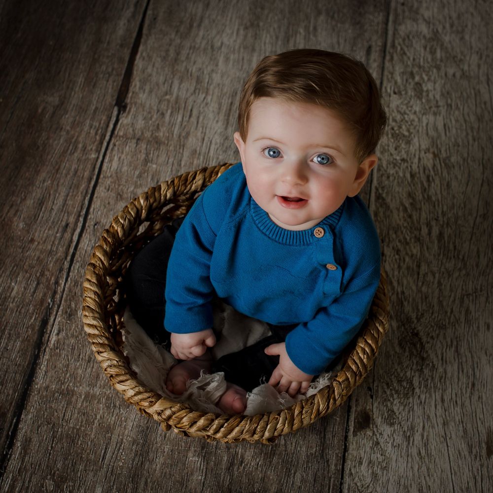 Baby in blue sweater sits in woven basket on dark wooden floor.