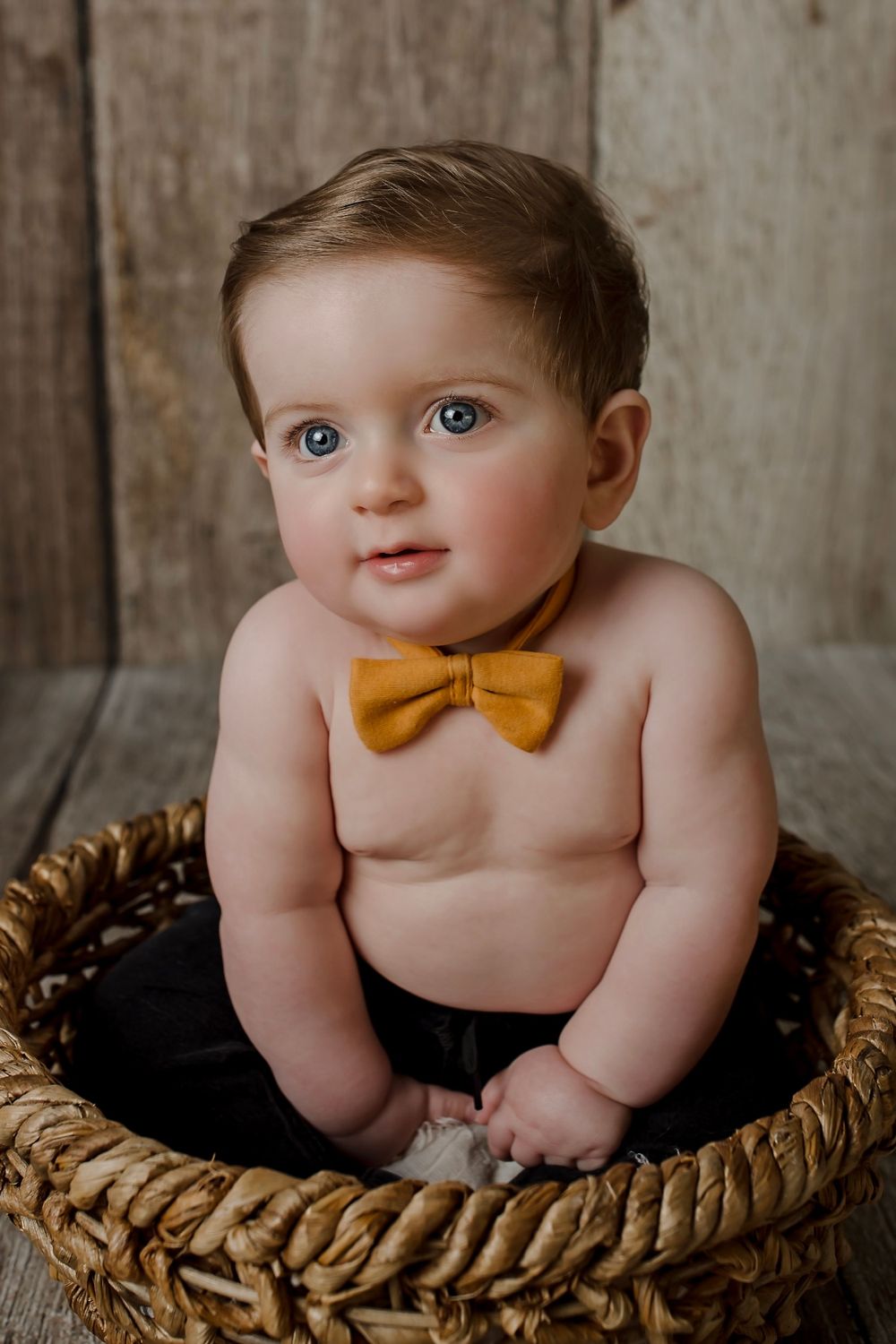 Baby wearing mustard bowtie poses in woven basket against rustic background.