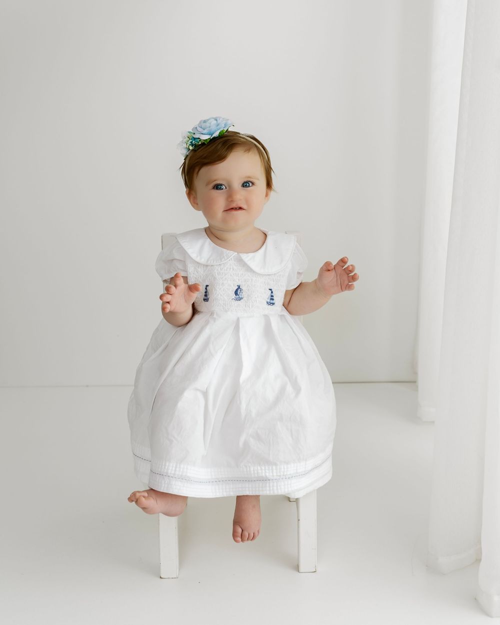 Baby in white dress and blue bow sits on small white chair against plain backdrop.