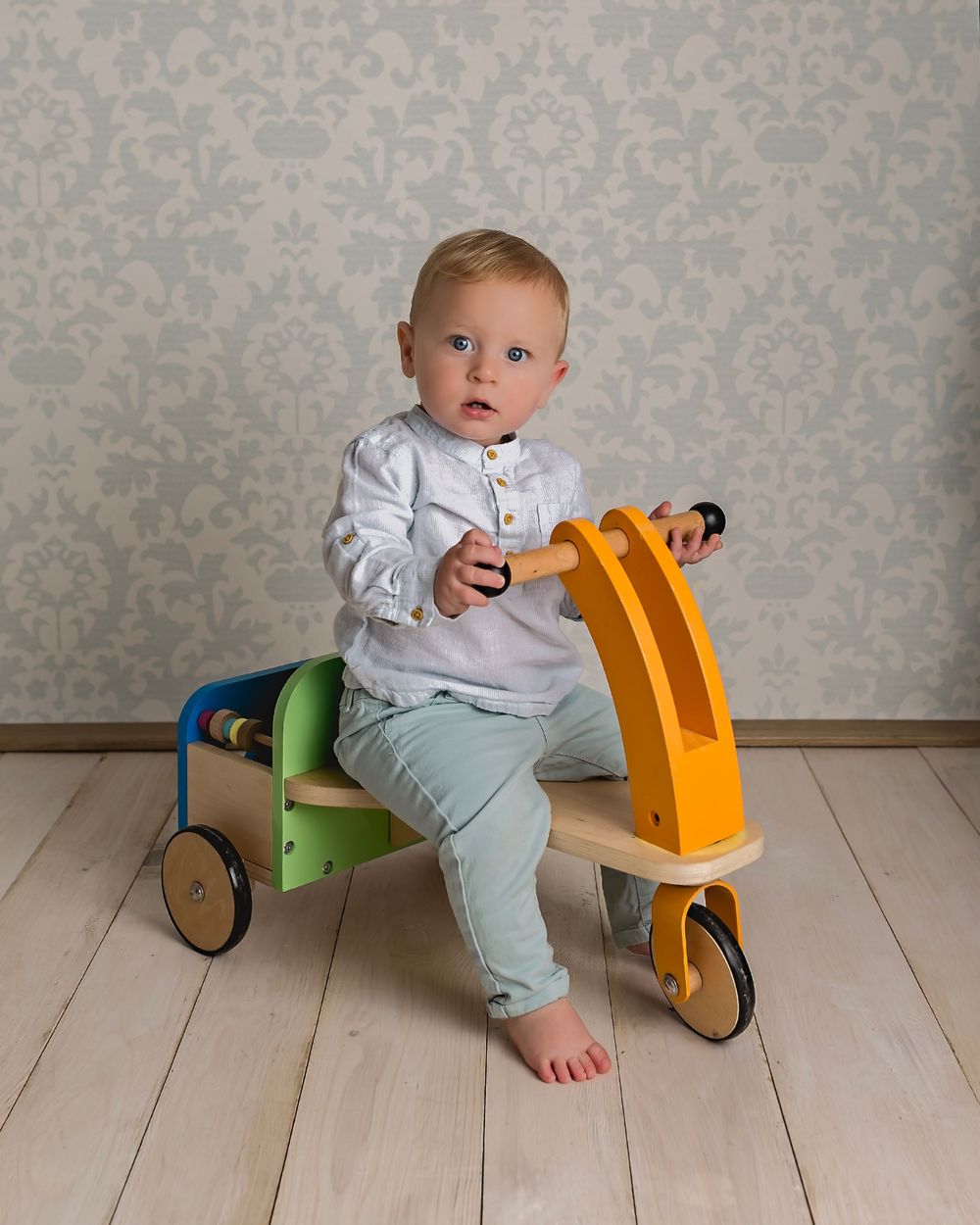 Baby rides colorful wooden push bike toy on light wooden flooring.