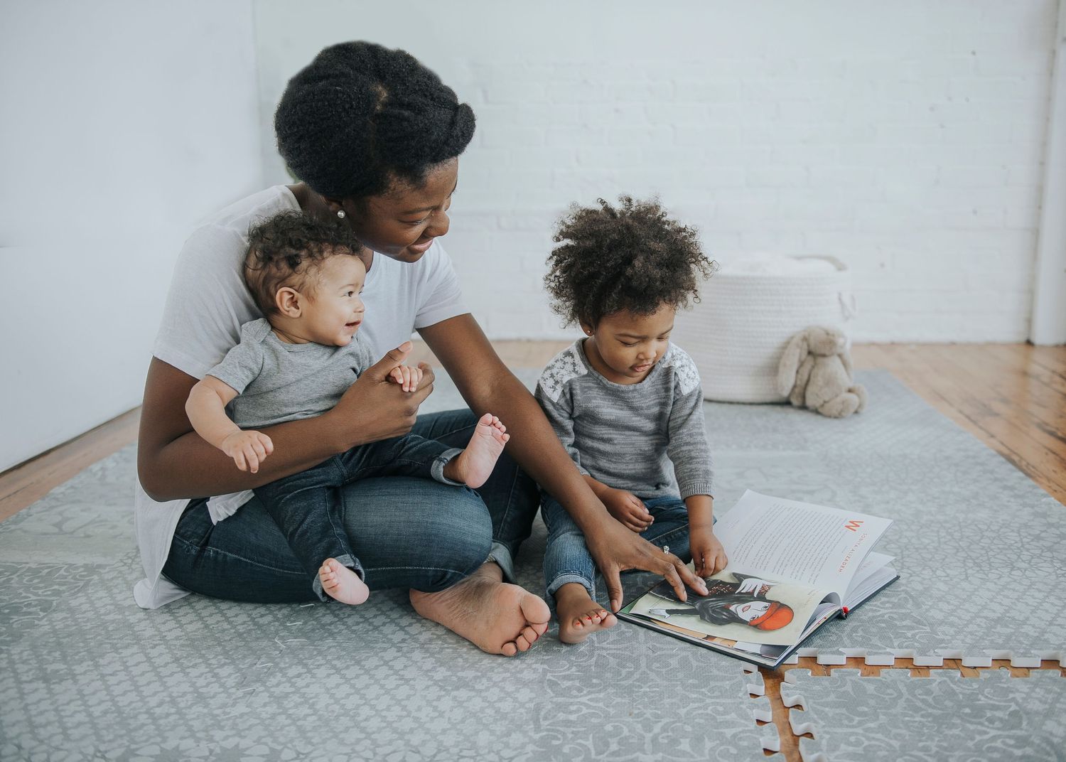 Parent reading a book with two small children on a gray floor mat at home.