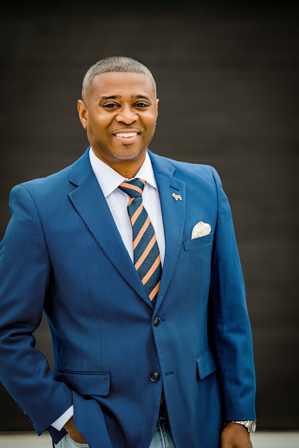 Professional portrait of man in blue suit with striped tie against dark background.