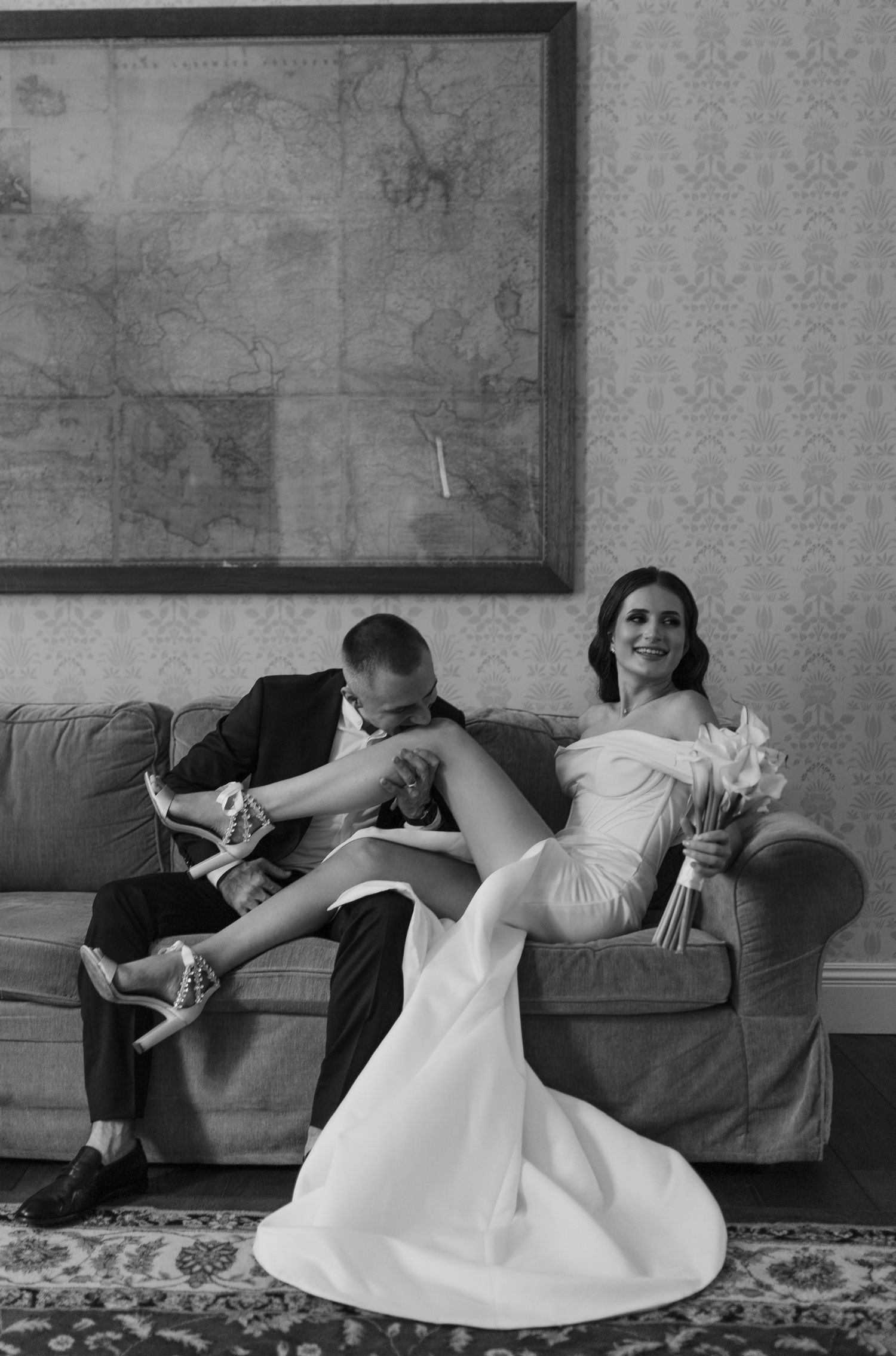 Black and white photo of couple lounging casually on sofa with floral artwork in background.