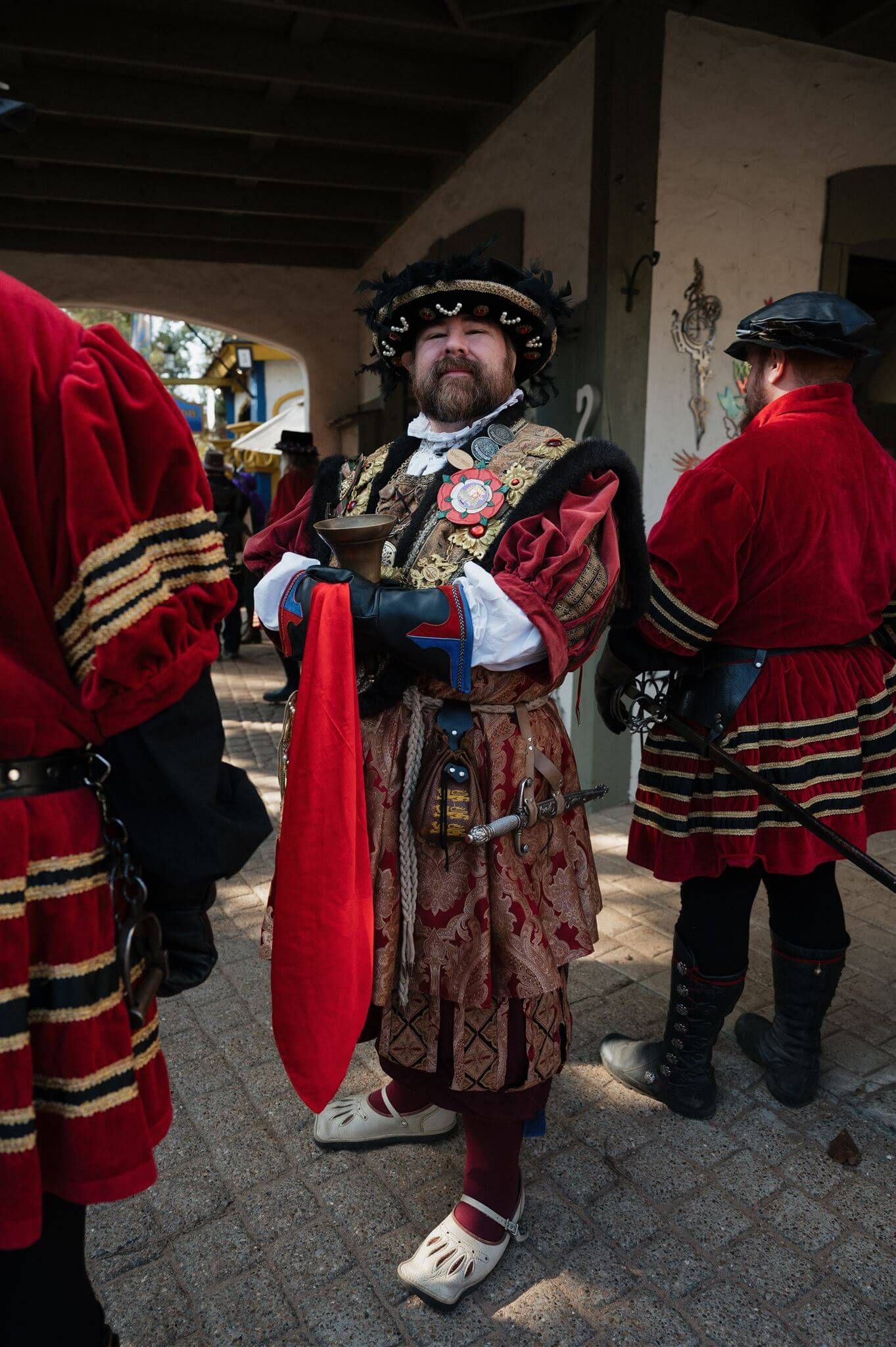 A performer in ornate Tudor-style costume with red and gold details stands at a Renaissance festival.