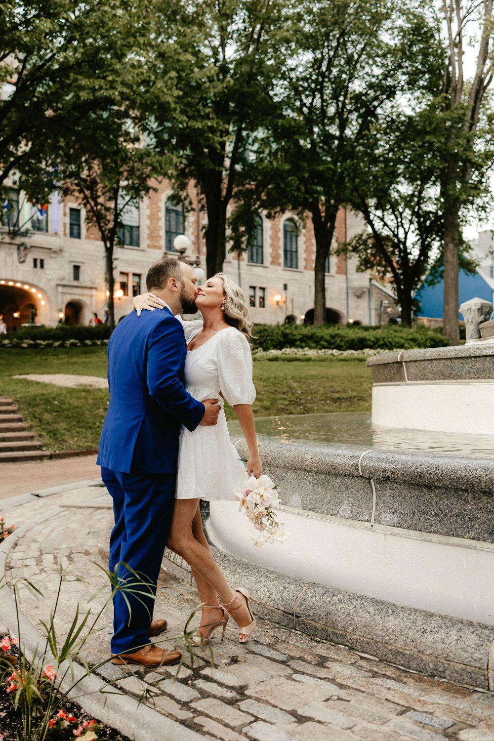 A couple embraces on stone steps near a grand historic building with a flower bouquet.