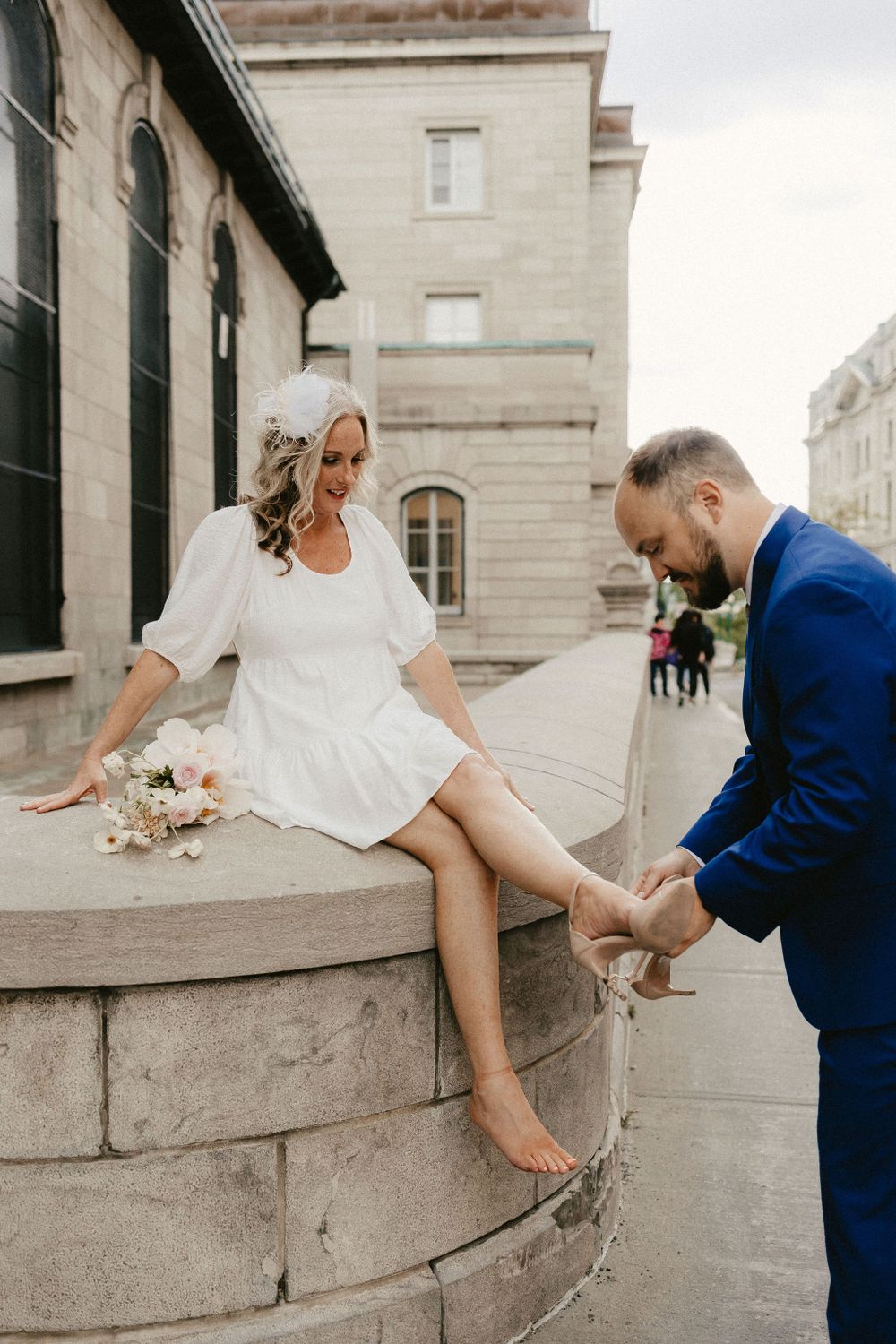 A couple shares a playful moment on a stone wall with a bouquet of flowers.
