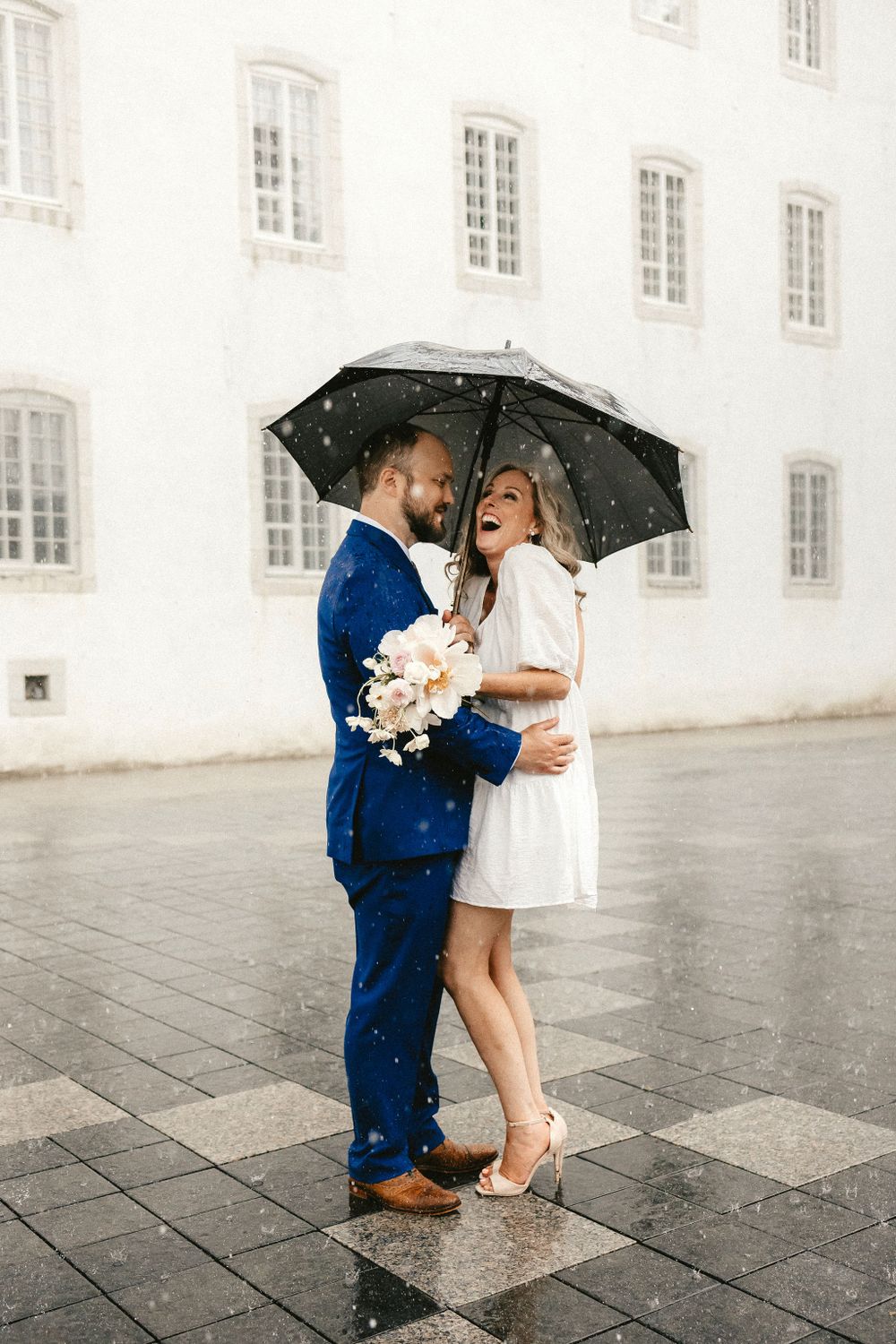 Couple shares intimate moment under black umbrella on rainy day, with white building and windows in background.