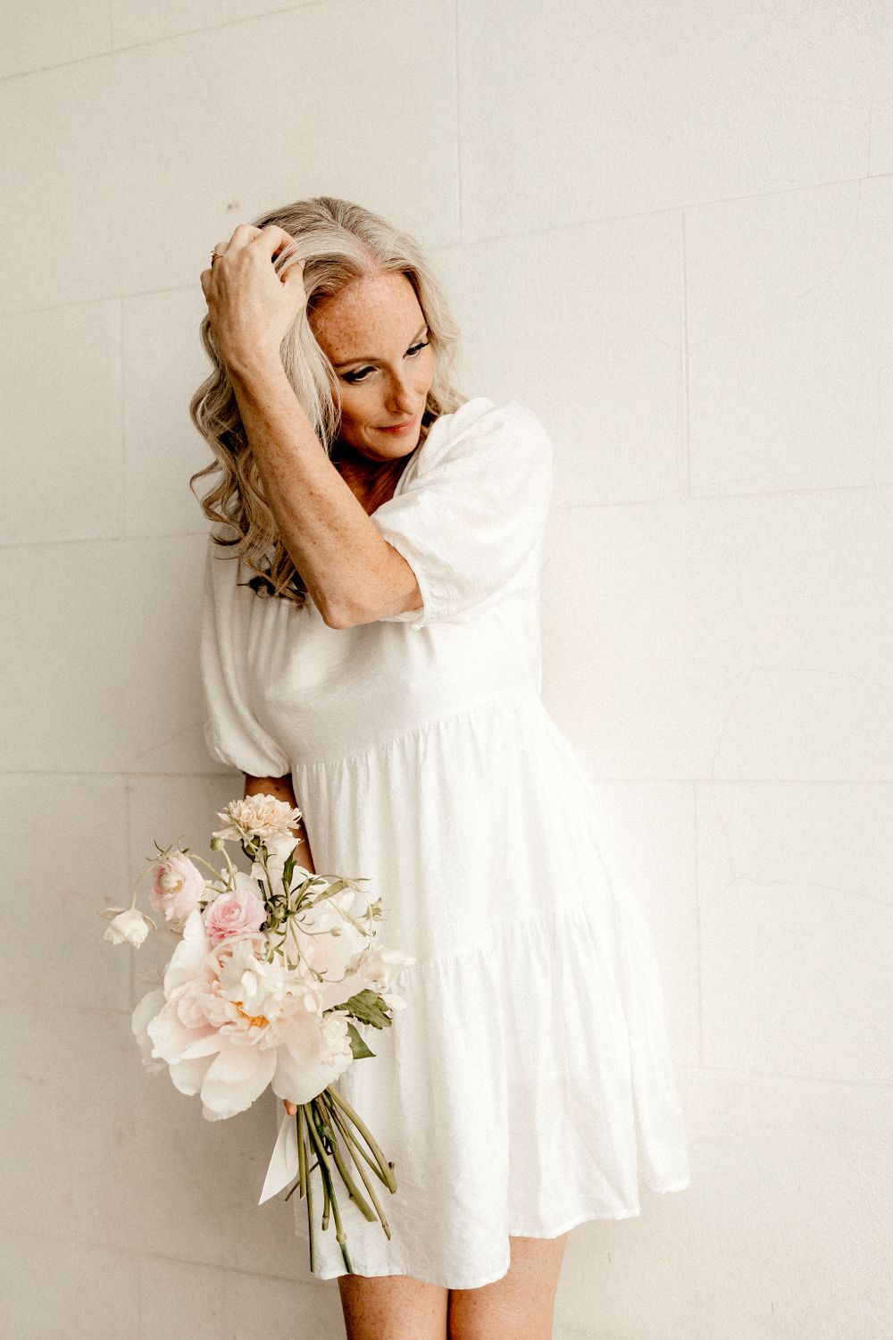 Woman in white dress holds delicate pink bouquet against neutral wall.