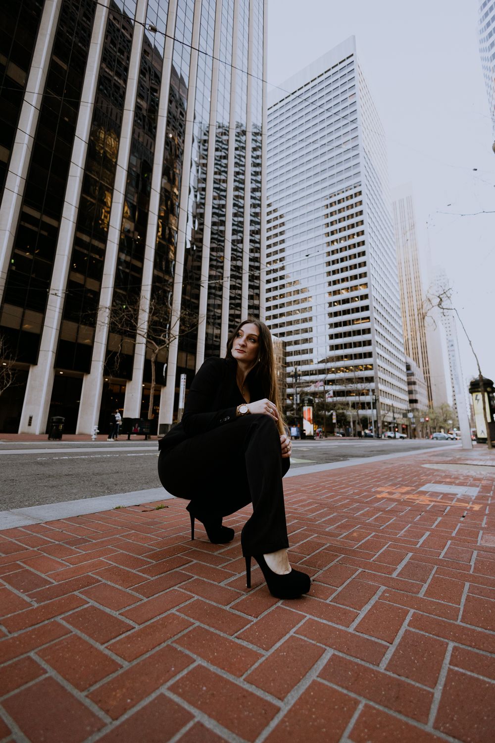 Stylish woman in all black crouches on brick pavement with downtown skyscrapers towering behind.