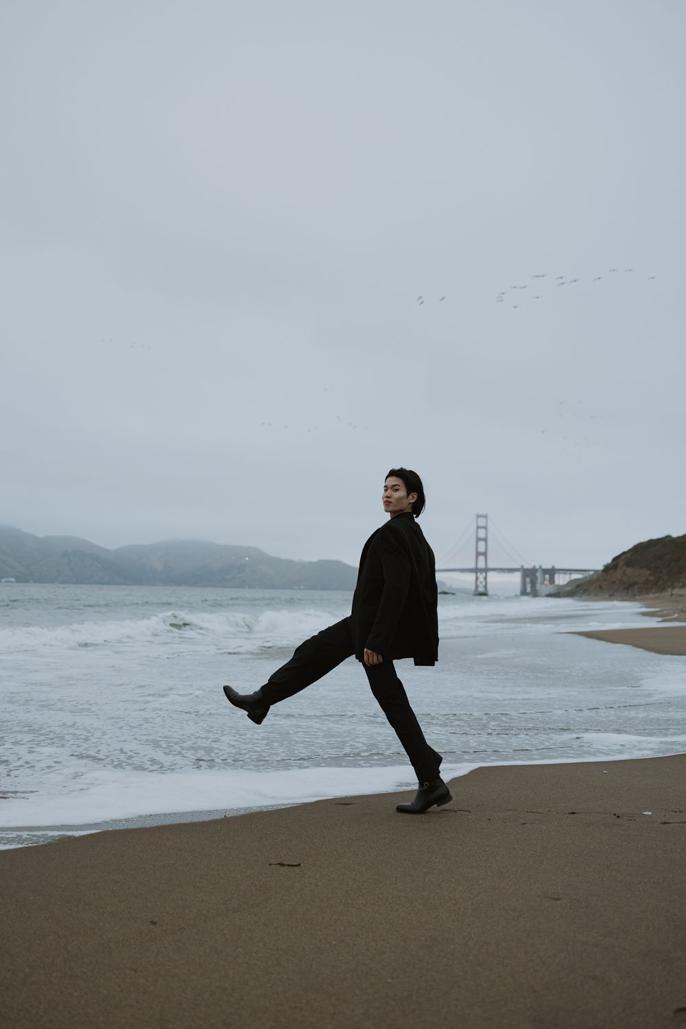 Artistic beach portrait with person kicking leg while Golden Gate Bridge visible in background.