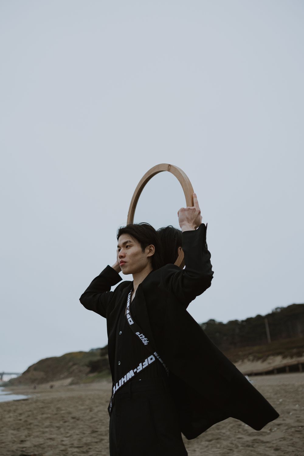 Person in black coat poses with circular prop against cloudy coastal backdrop.