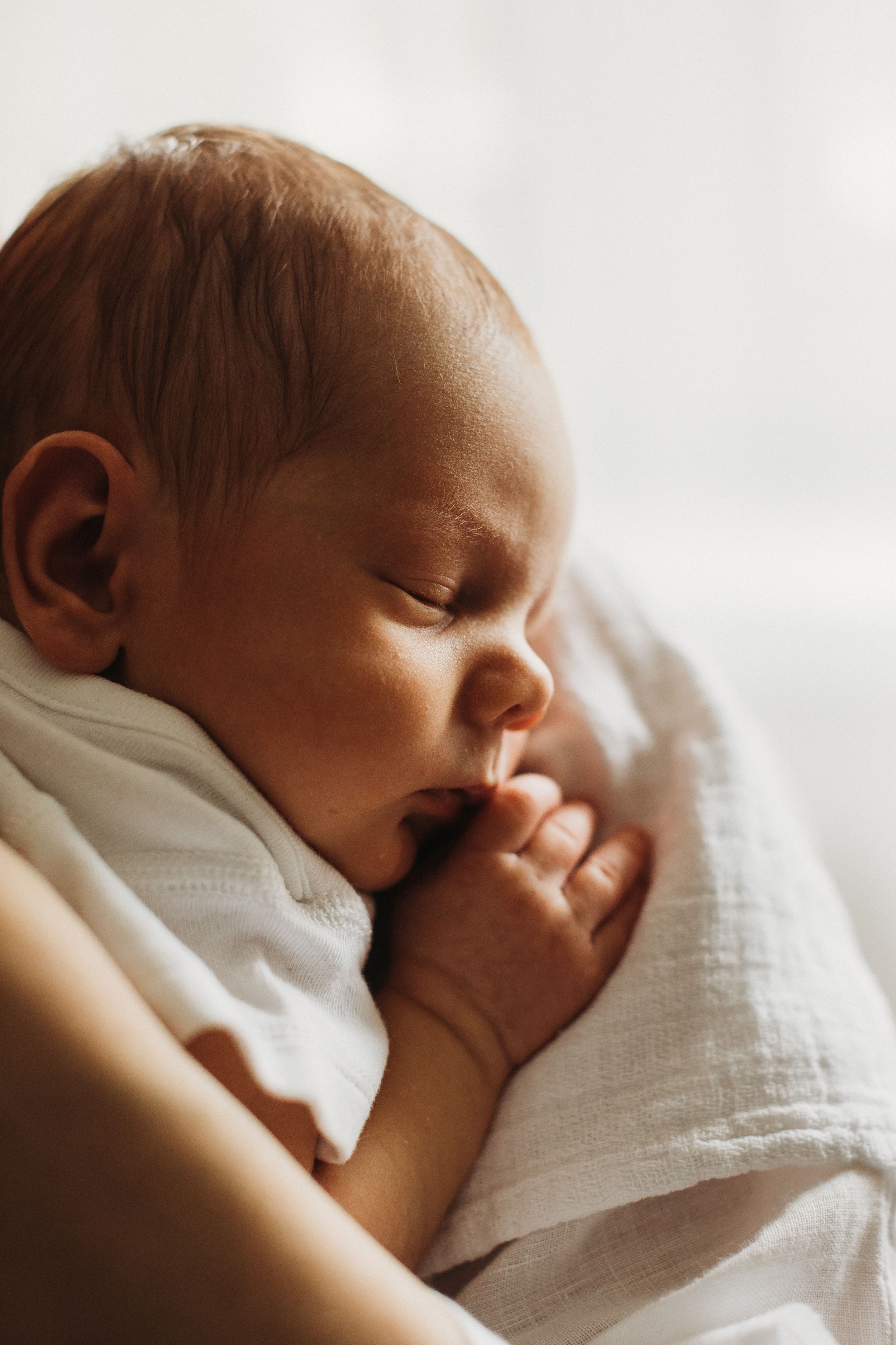 Peaceful sleeping newborn baby wrapped in white muslin cloth sucking tiny fingers.