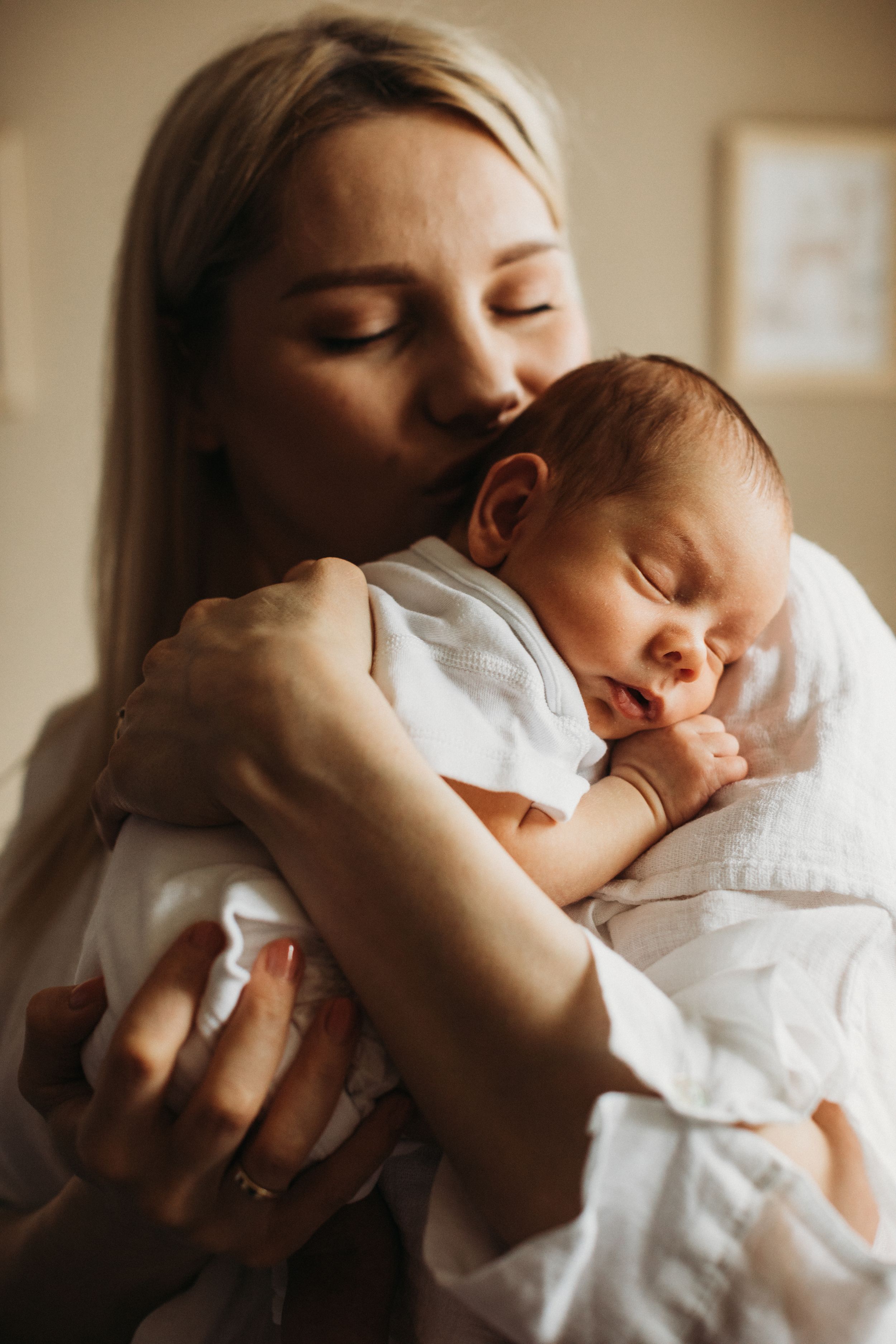 Blonde mother gently kissing sleeping newborn baby wrapped in white blanket indoors.