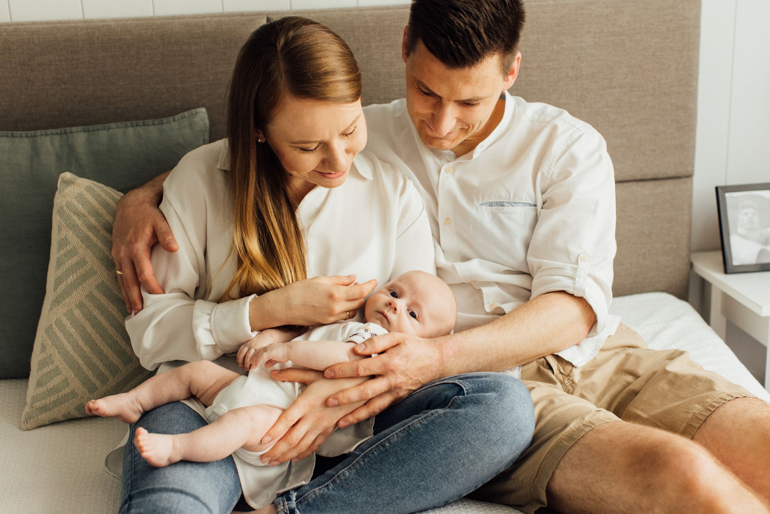 Young couple sitting on bed smiling while holding their newborn baby.