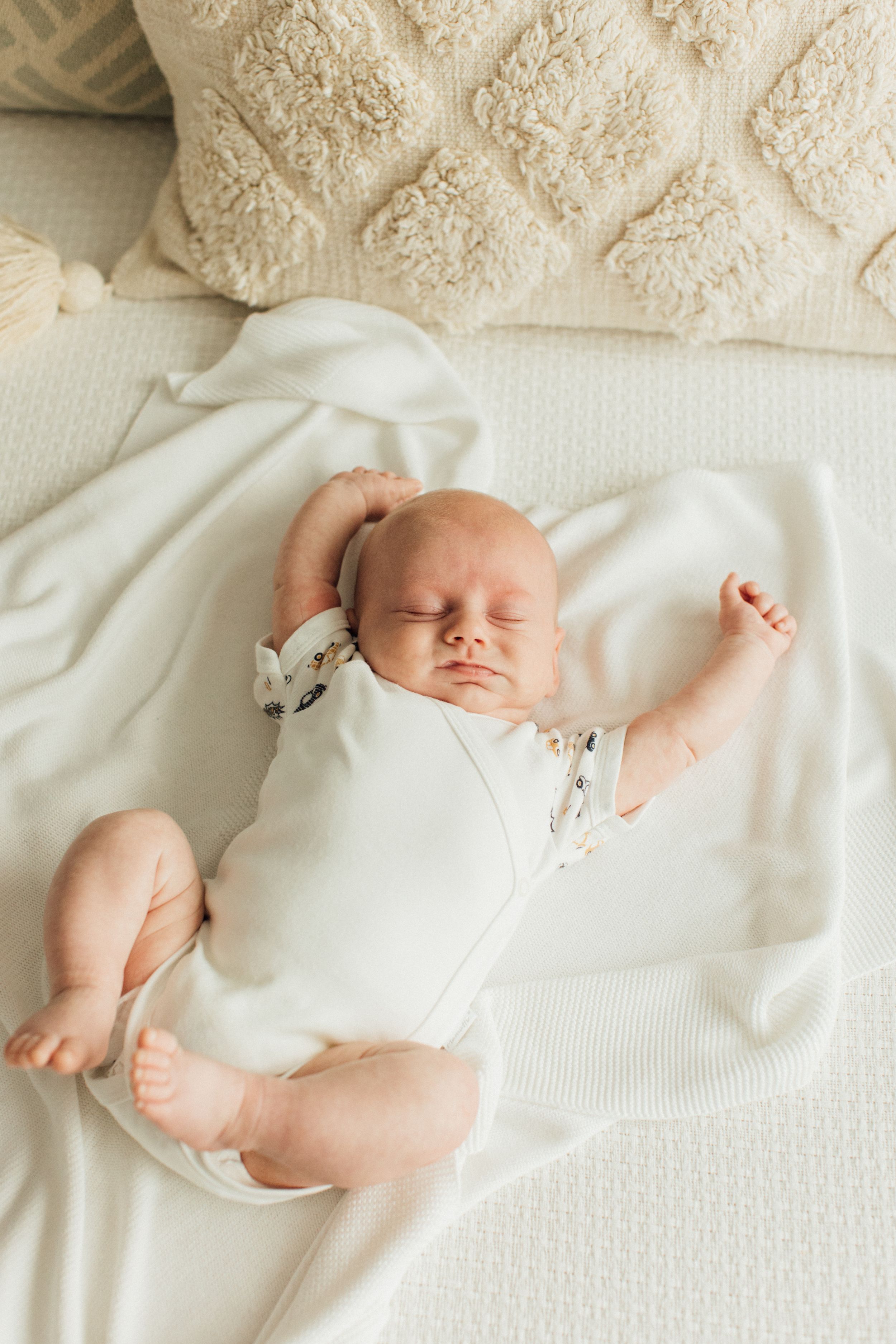 Newborn baby sleeping on white blanket surrounded by cream pillows in bright nursery.