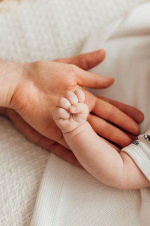 Tiny newborn baby hand resting on adult palm on white knit blanket.
