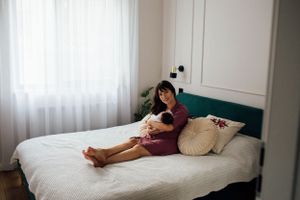 Mother in a burgundy dress sitting on a white bed cradling her sleeping newborn baby.