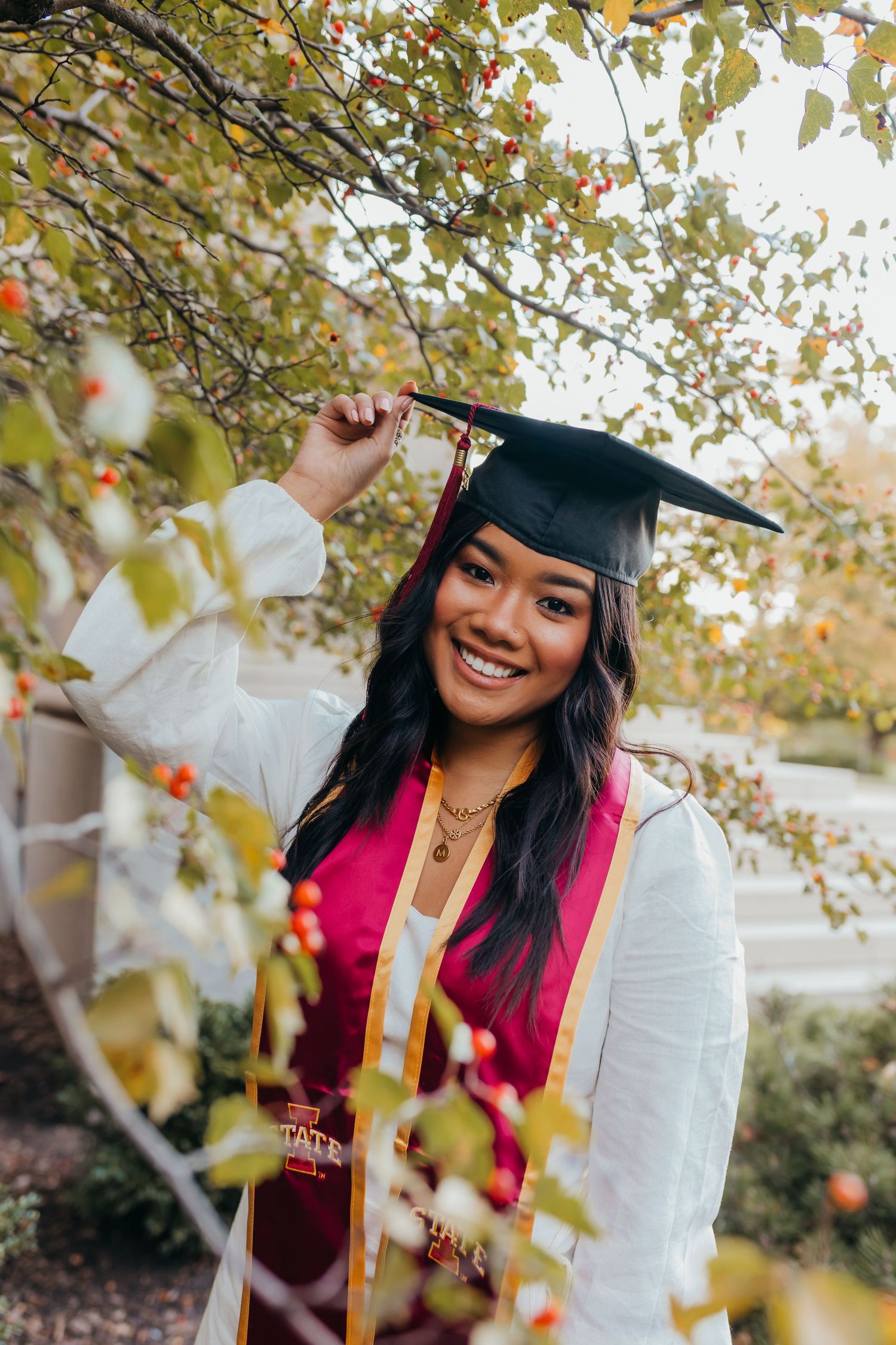 Graduate in cap and gown smiling outdoors surrounded by autumn foliage.