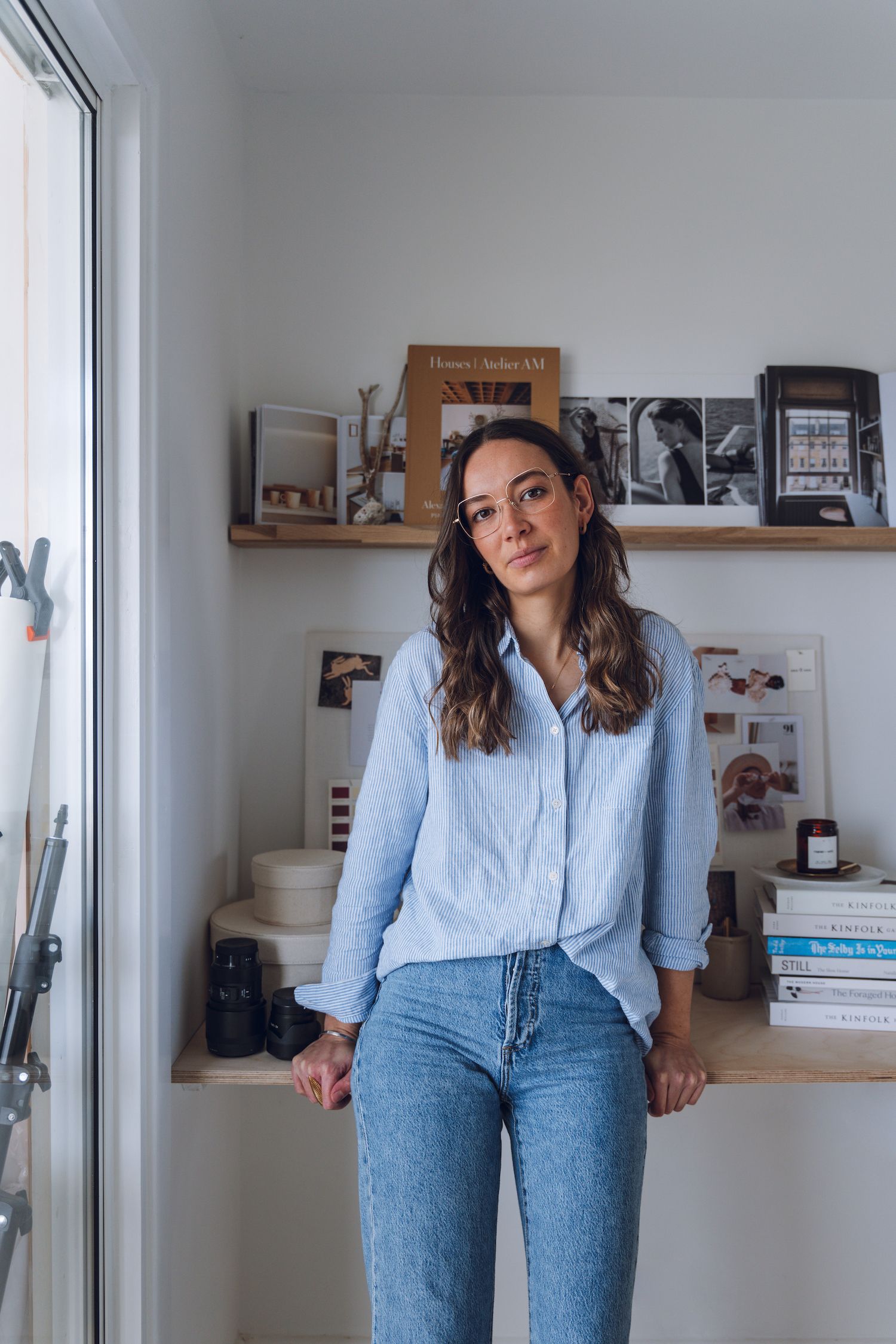 Woman in light blue shirt and jeans poses casually against white wall with floating shelves.