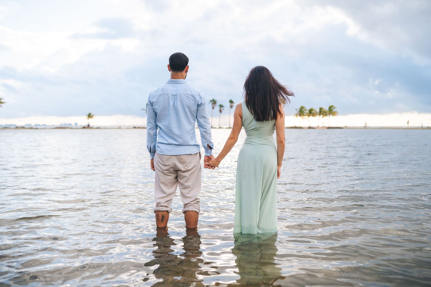 Couple holding hands while standing in shallow water at beach.